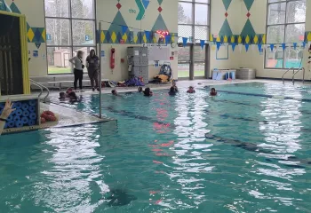 Kids participate in a swim lesson; instructors watch from the pool's edge.
