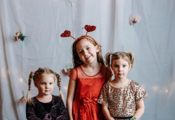 Three girls in party dresses pose for a photo. One wears a red dress with heart-shaped headbands, another a sparkly gold dress, and the youngest a dark burgundy dress.
