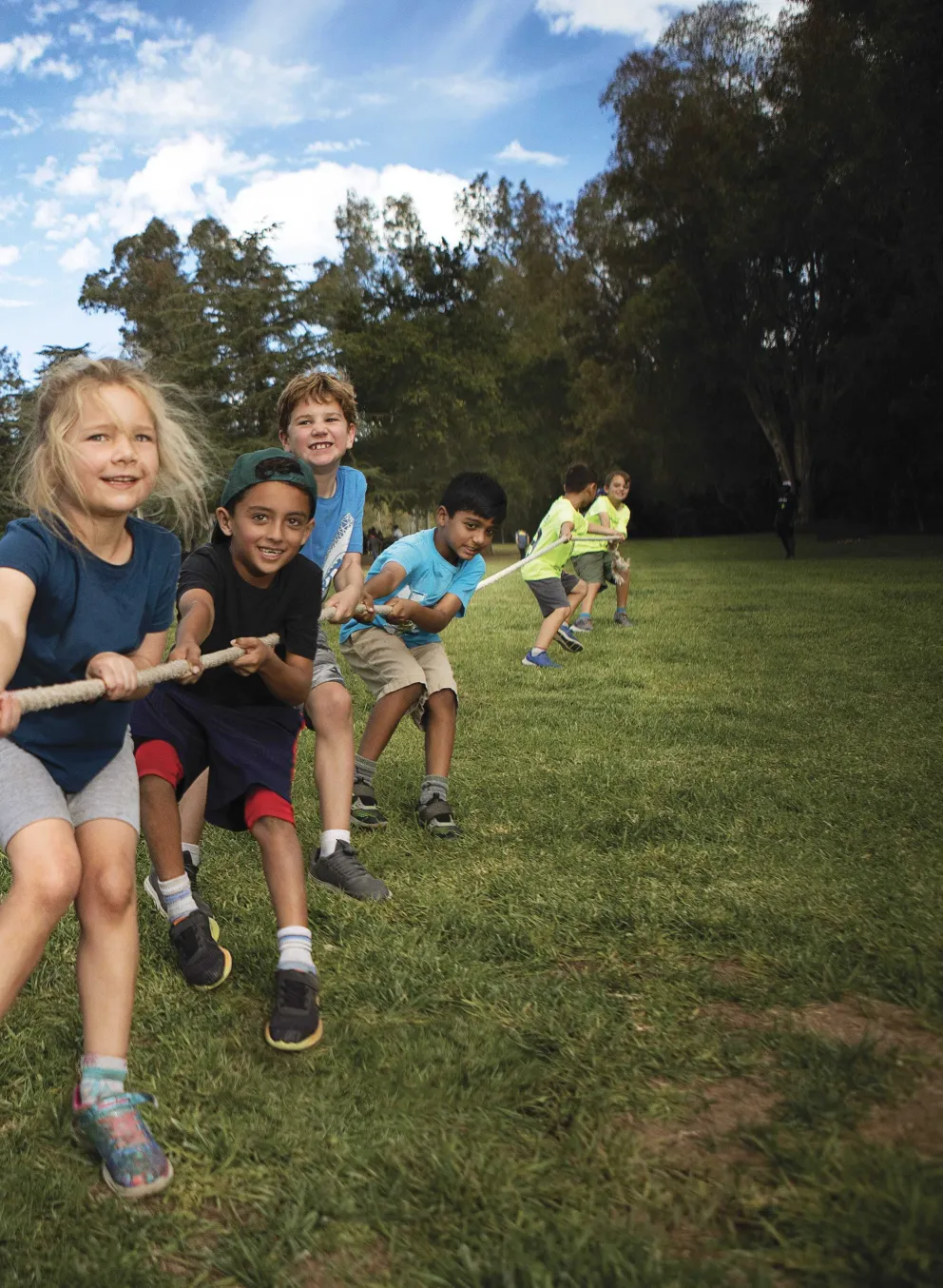 Kids joyfully play tug-of-war on a grassy field, under a bright blue sky.  Smiling children pull a thick rope, their faces full of energy and excitement.