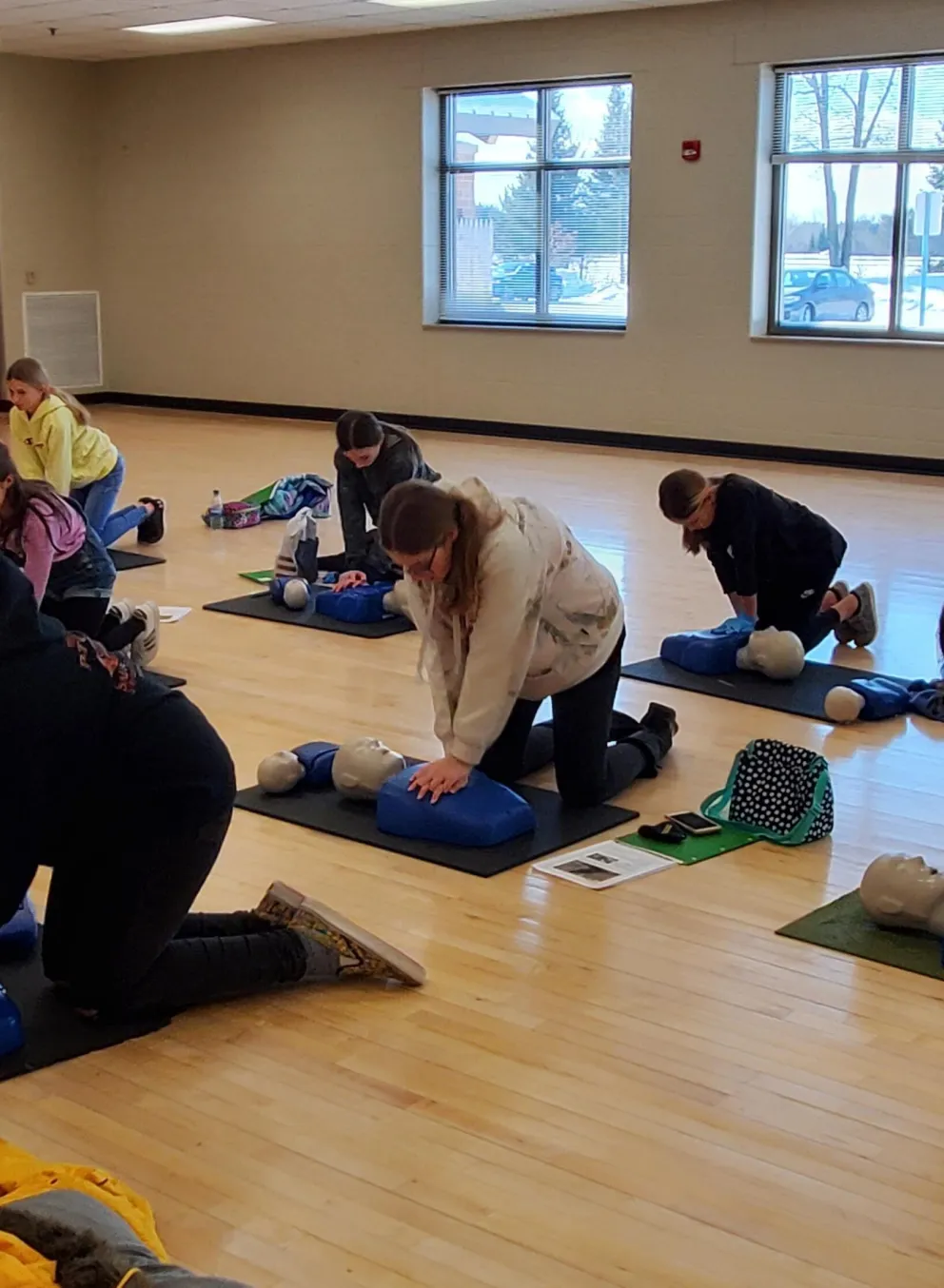 Several young adults kneel on mats, practicing CPR on training dummies in a gym.  Exercise balls are visible in the background.