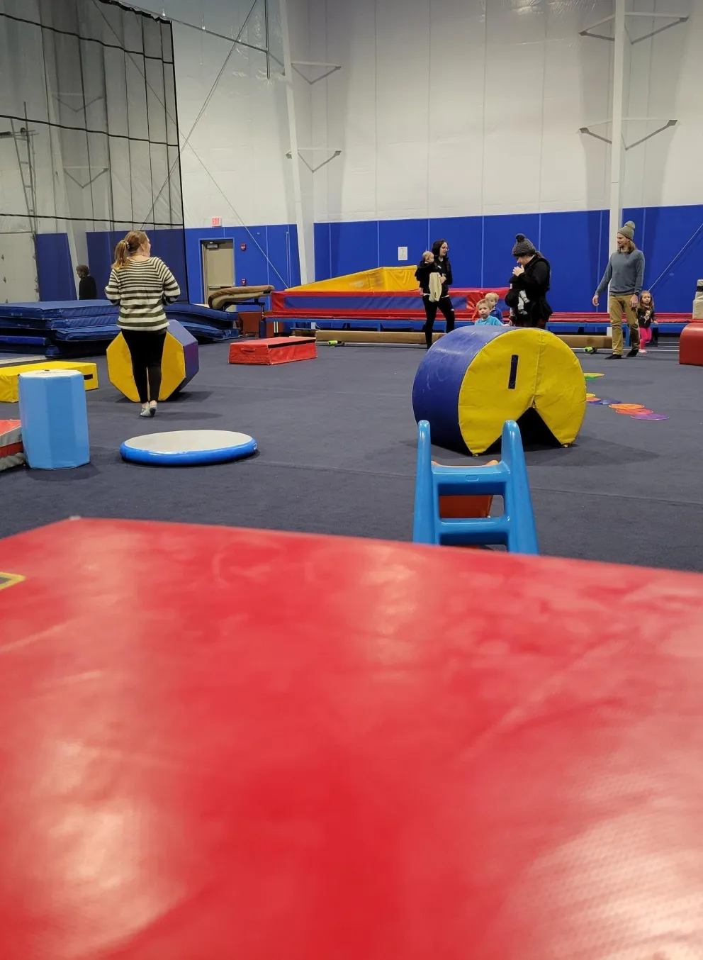 Kids' gymnastics class in session.  Toddlers and adults are present. Colorful mats, blocks, and other equipment are scattered around a large gym floor. A little girl is playing near some mats.
