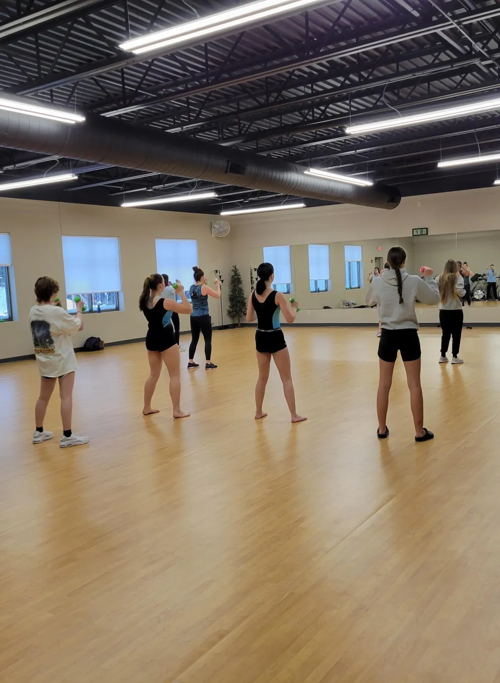 Young women in athletic wear participate in a fitness class, some holding light weights, in a bright, spacious room with wood floors and large windows.
