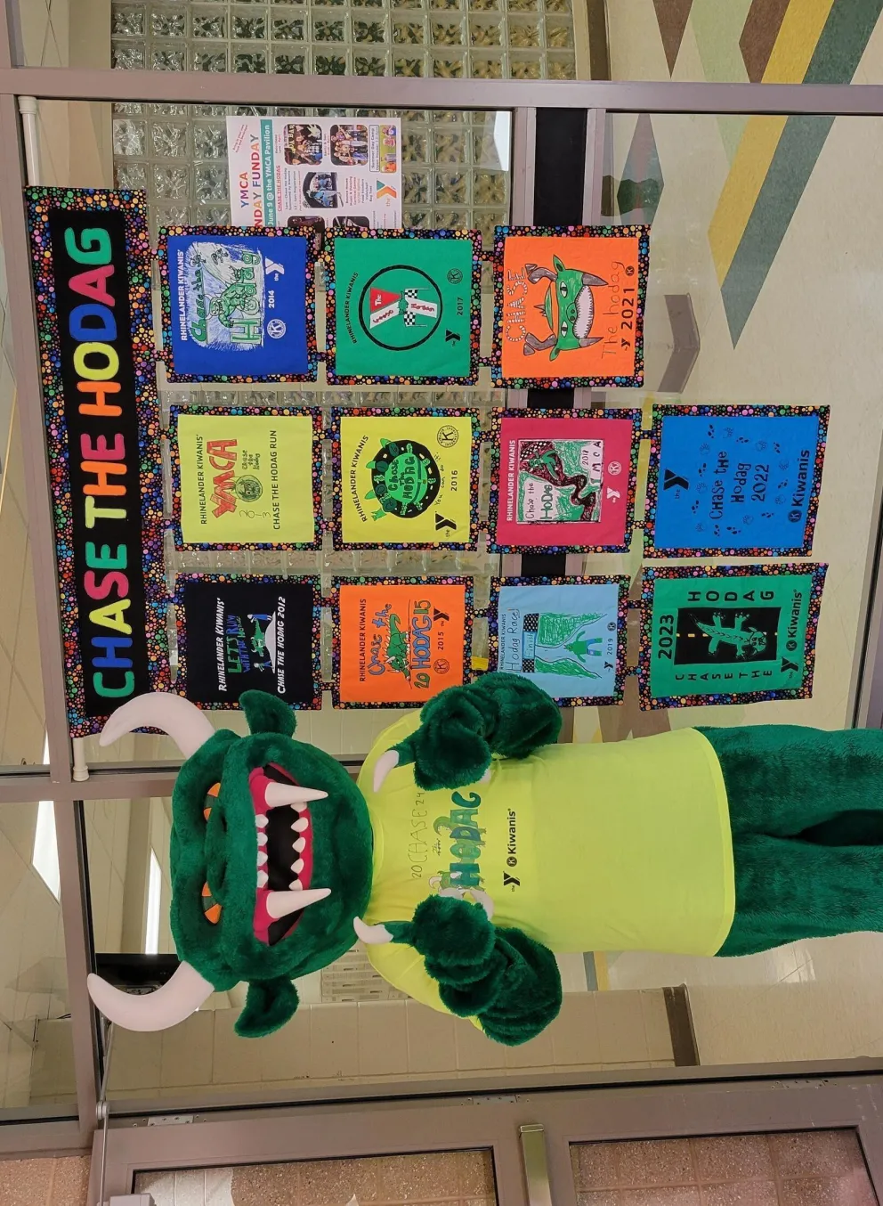 Green Hodag mascot in lime green shirt stands by colorful event posters;  "Chase the Hodag" banner above.