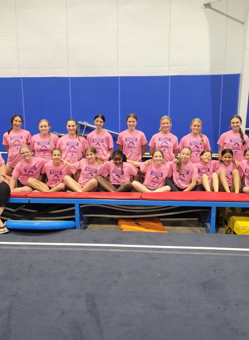A group of girls in pink t-shirts sits on a padded gymnastics platform, two adult women flank them.  A trampoline is behind them.