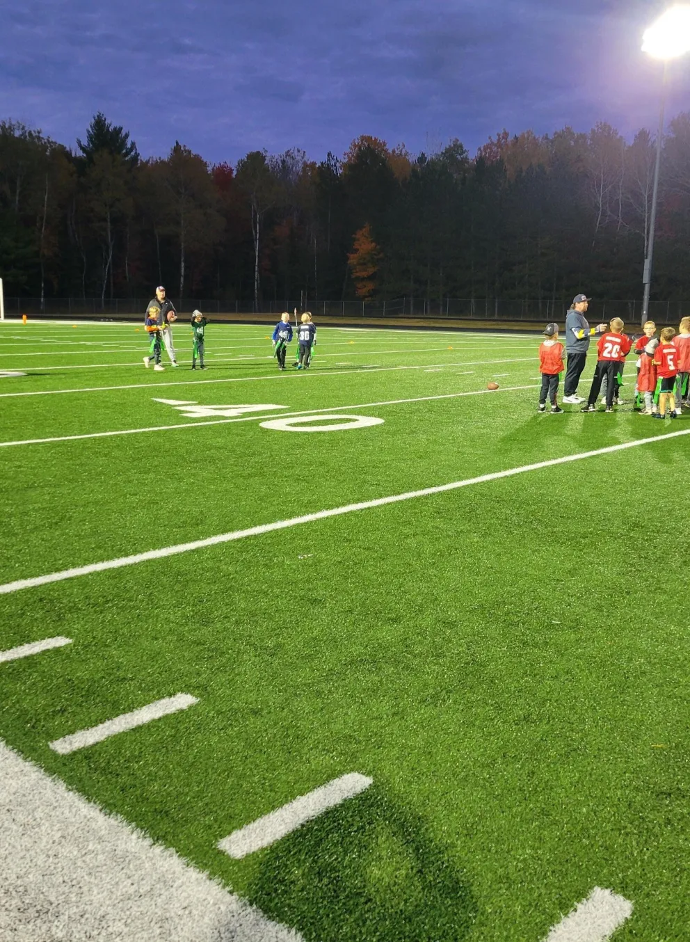 Kids in red jerseys participate in a football practice on a well-lit field, with coaches providing instruction under the evening sky.  A few other kids and adults are nearby.