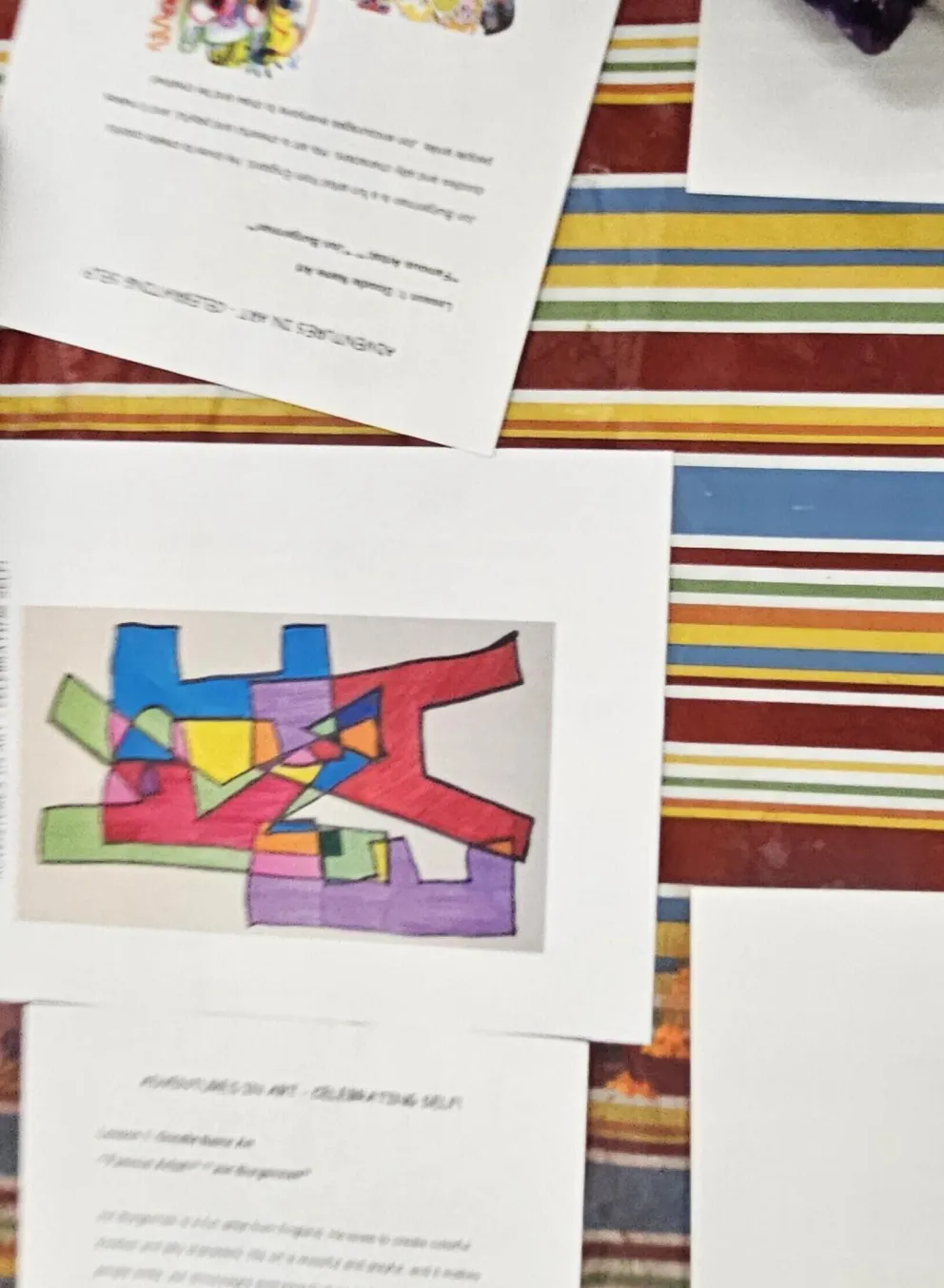 Colorful markers in a container, papers with instructions and a child's abstract art piece on a striped table.