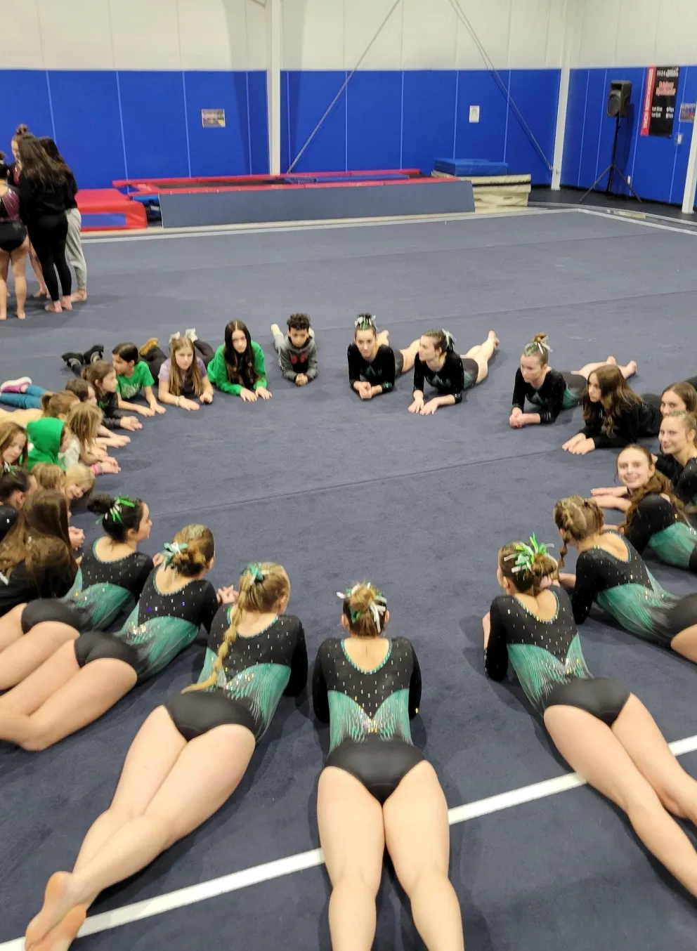 Gymnasts in teal leotards lie on the floor in a circle facing inward, several coaches and a group of other gymnasts stand in the background.