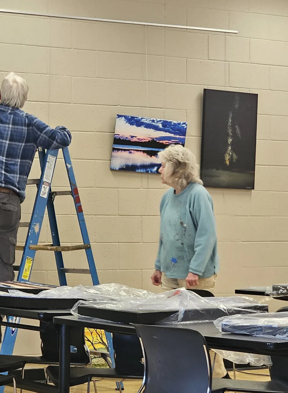 Two adults hang artwork on a wall; one uses a stepladder.  Several framed landscape photos are visible, along with tables covered in plastic-wrapped items.
