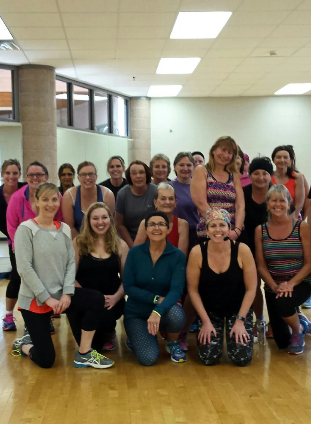 A large group of women and one man pose in a gym; many are kneeling.  They appear to be participating in a fitness class.