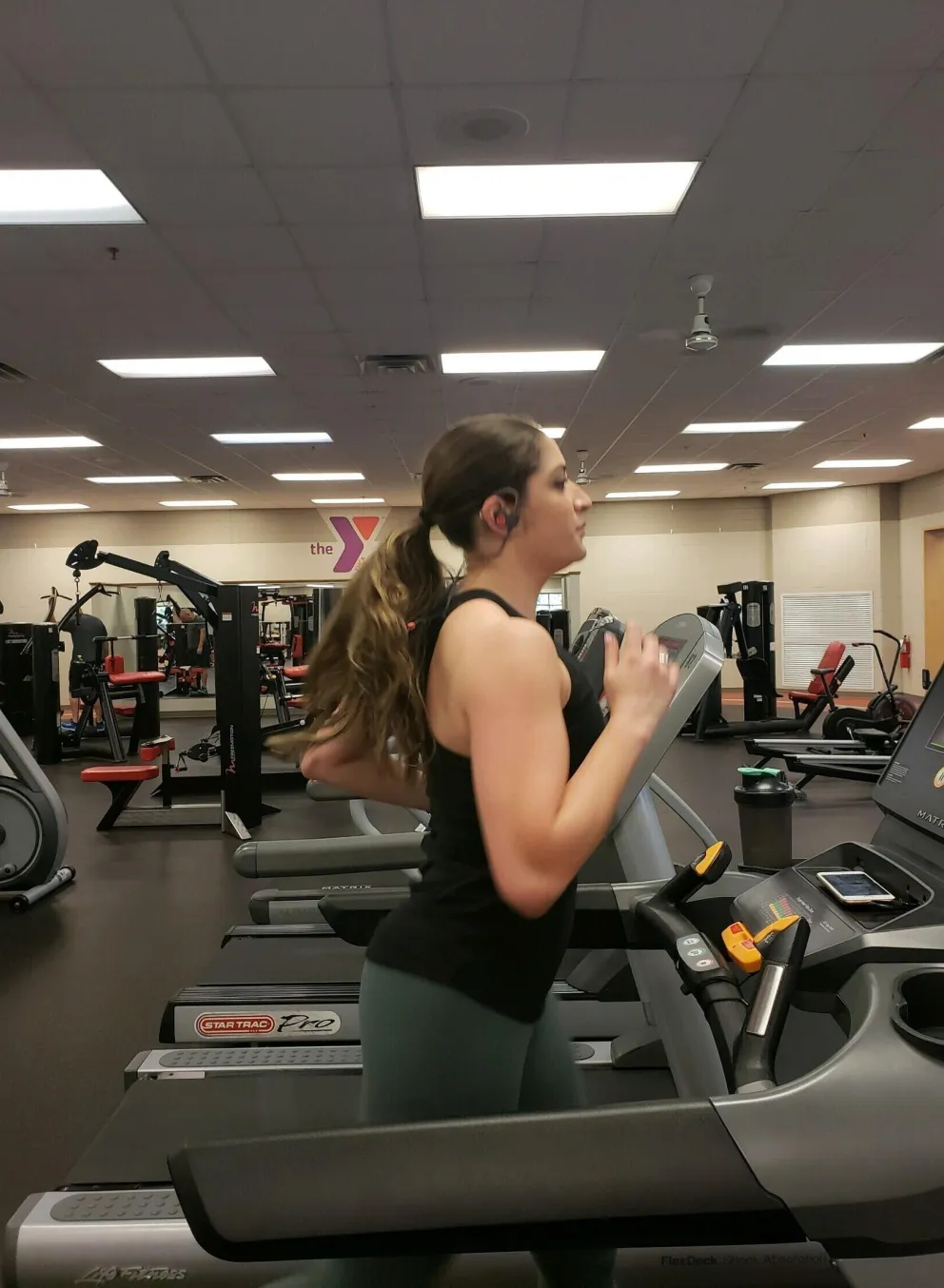 A woman with long brown hair runs on a treadmill in a fitness center; other exercise equipment is visible in the background.