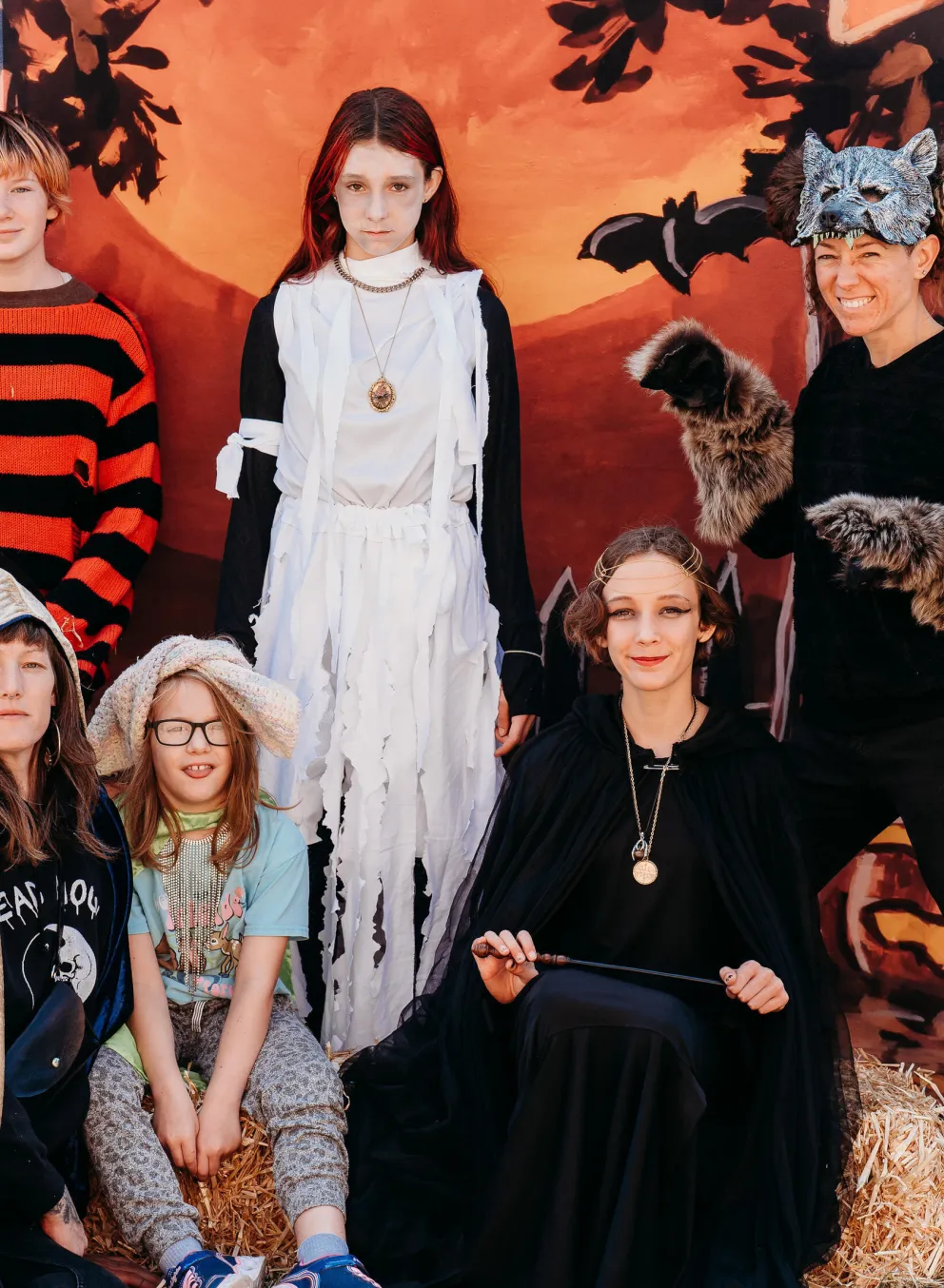 Kids in Halloween costumes pose for a photo; a girl dressed as a ghost, others in fantasy attire,  against a painted backdrop.