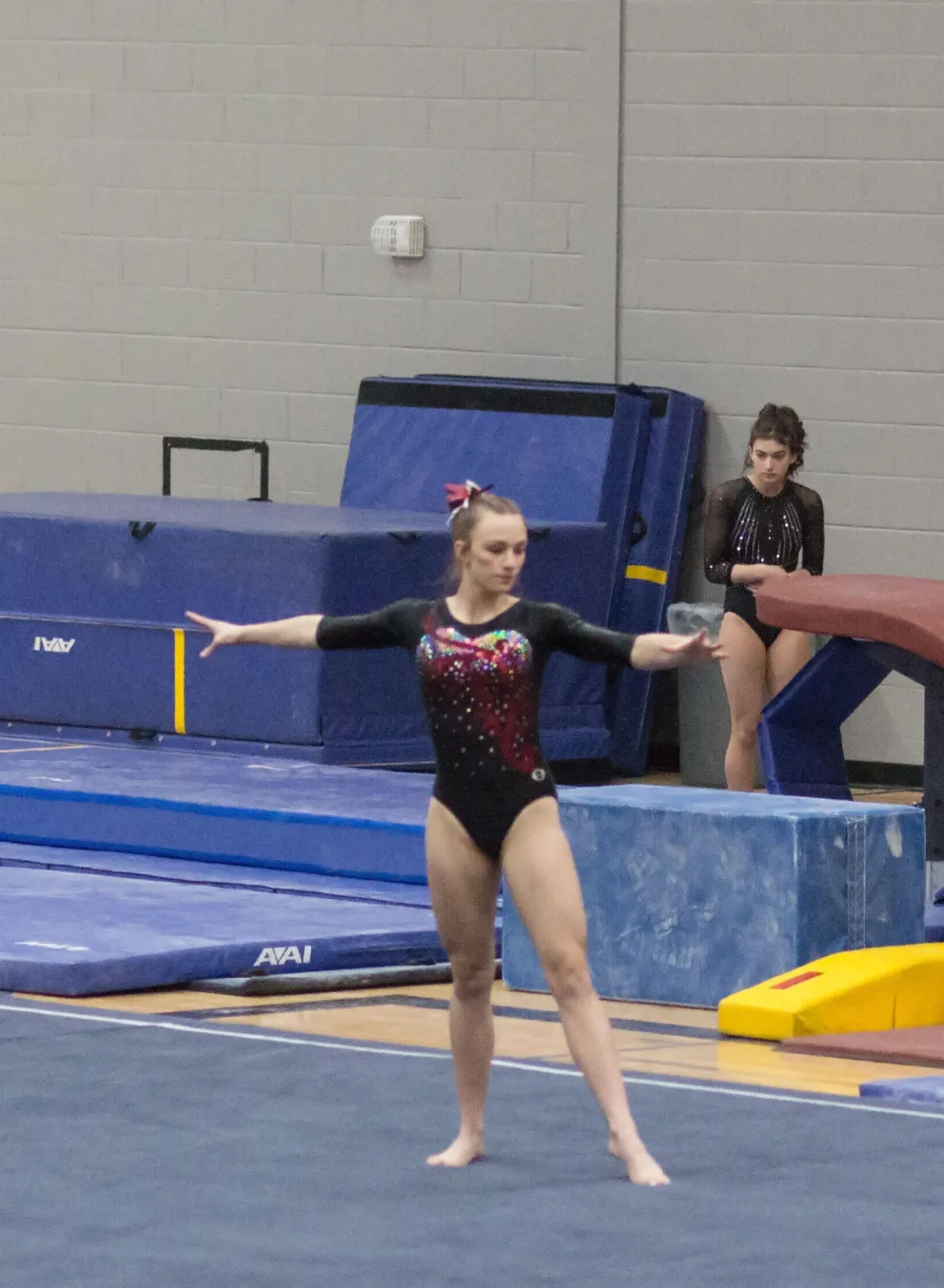 A young gymnast in a black and red leotard performs a floor routine; other gymnasts watch nearby.