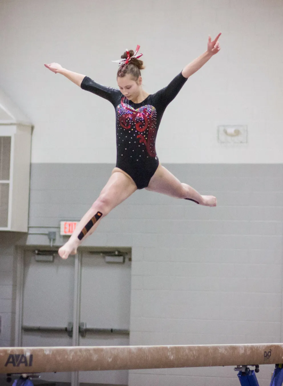 A young gymnast in a black leotard with red accents performs a graceful leap over a balance beam.  Spectators watch from the sidelines.