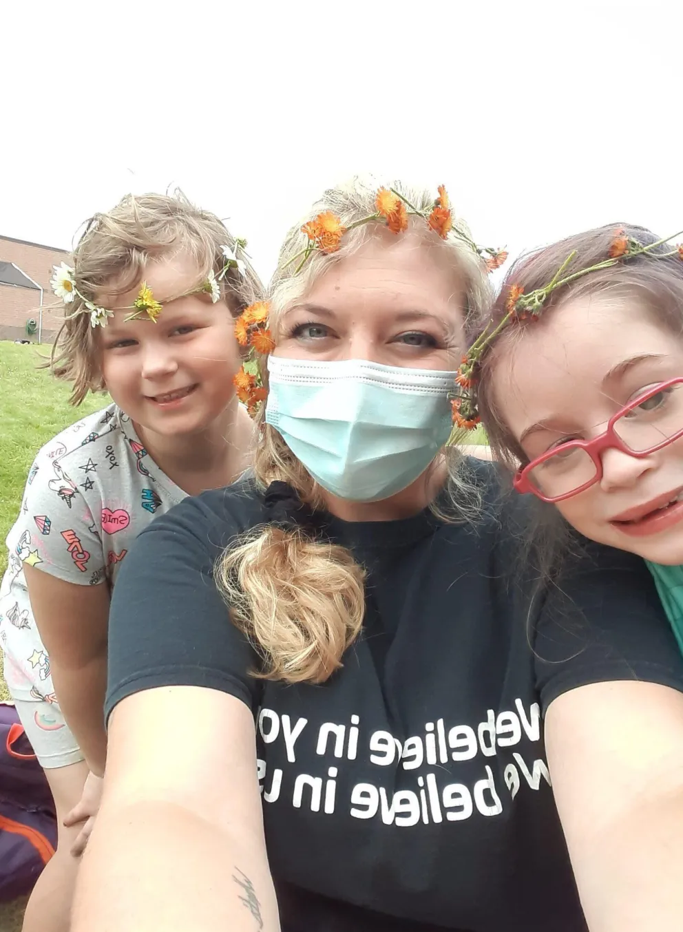 Smiling children and a counselor wear flower crowns outdoors.  The counselor wears a mask and a black t-shirt with mirrored text.  A fun summer camp moment.