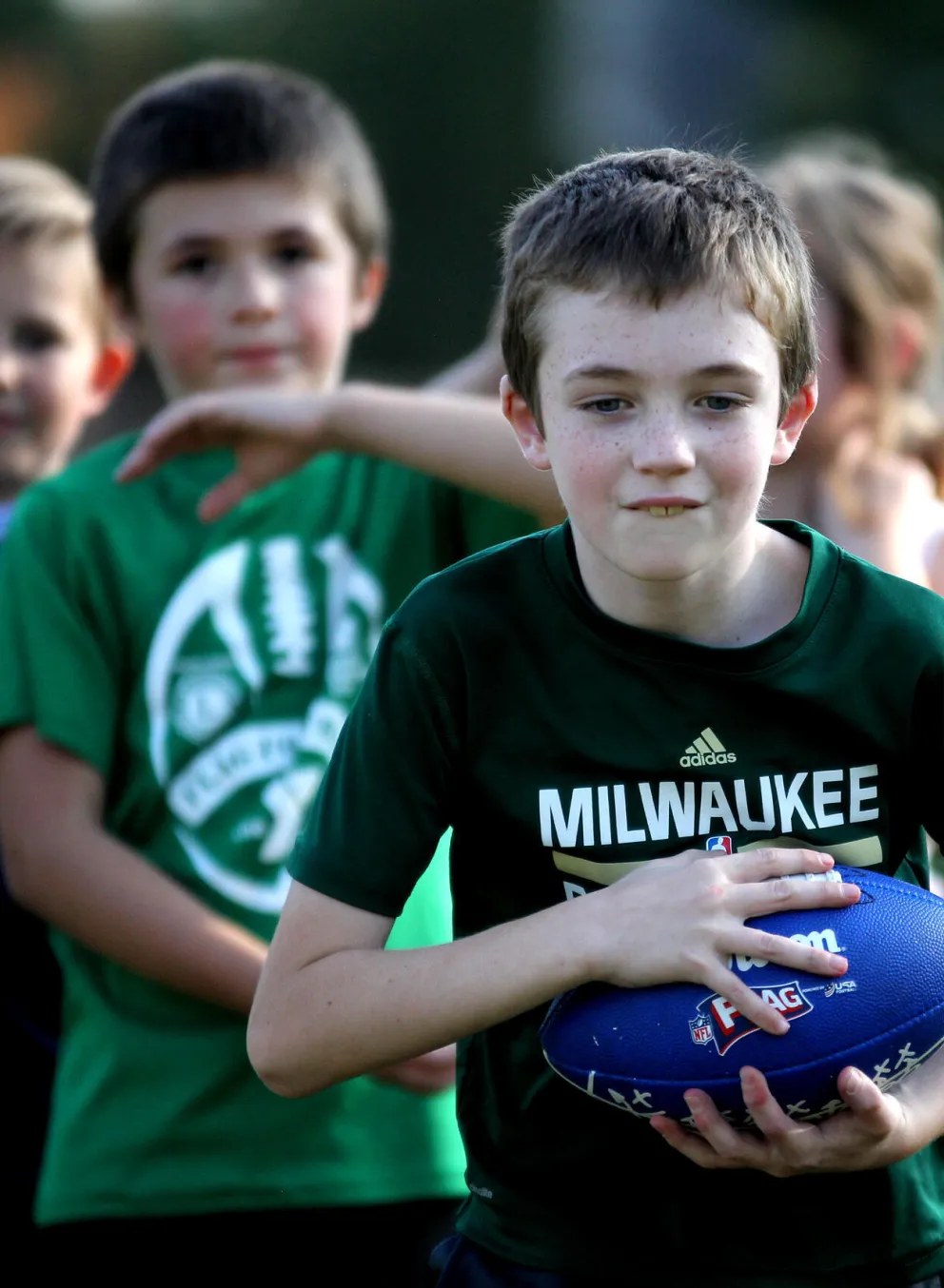 Kids in green shirts participate in a youth sports program; one boy holds a blue football.