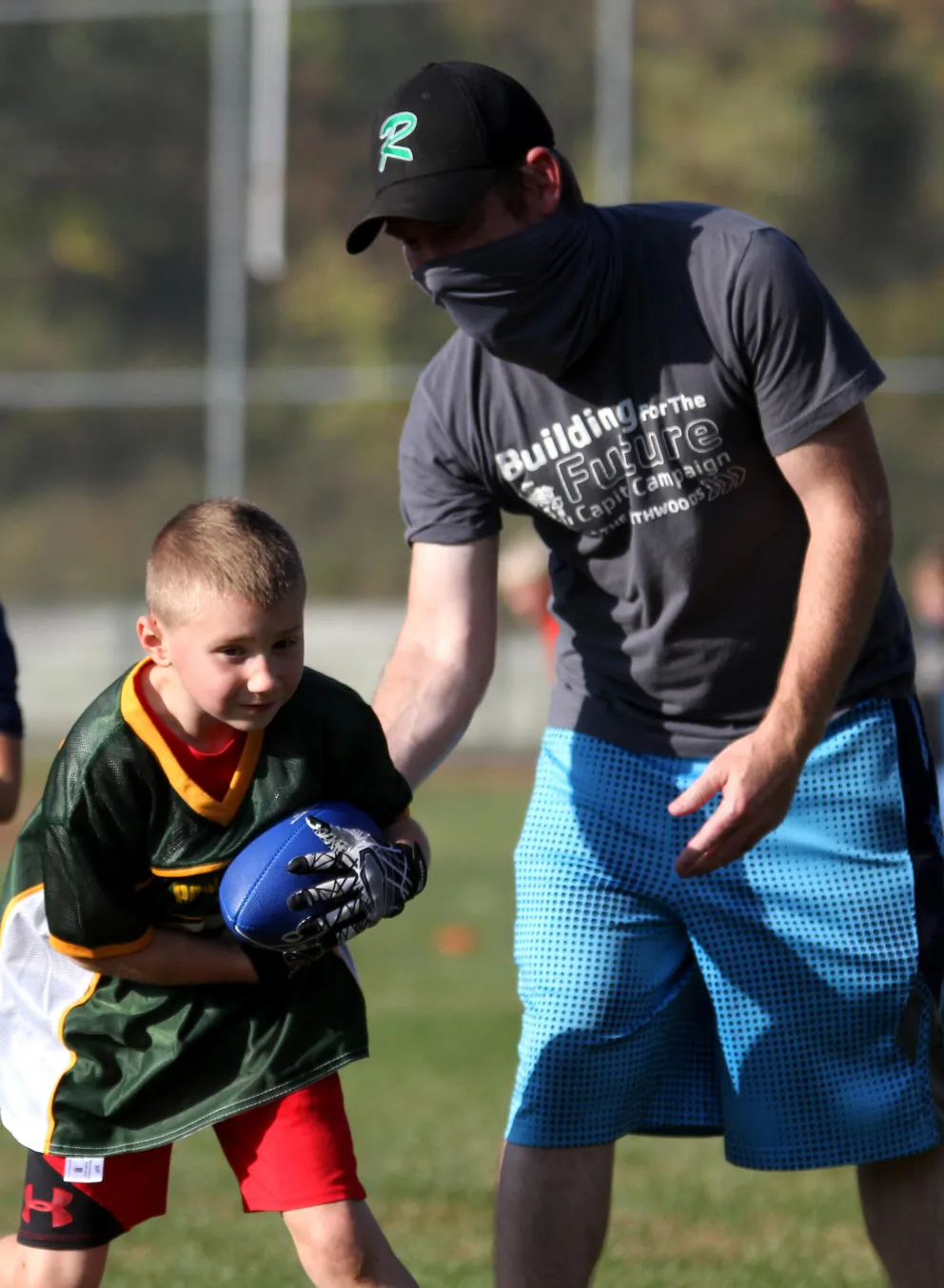 A young boy in a football uniform grips a ball, receiving coaching from a man wearing a face covering and a gray "Building for the Future" campaign shirt.  They're on a grassy field.