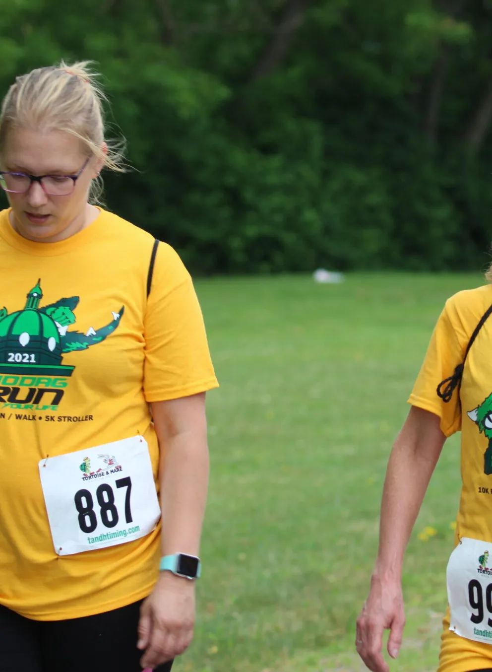Two women in yellow t-shirts, bearing the 2021 Holcomb Run logo, walk outdoors.  Their race bib numbers are 887 and 991.