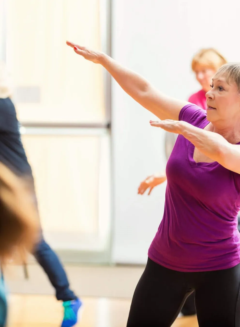 Women of varying ages participate in a fitness class, gracefully moving their arms and bodies in unison.  One woman in a purple shirt is prominently featured, her arms outstretched.