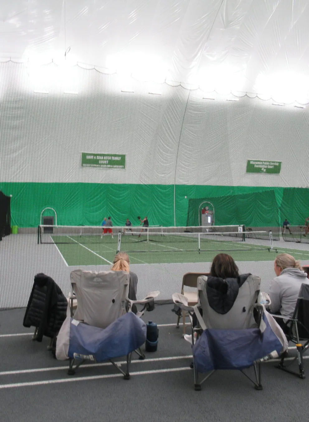 Indoor tennis court with spectators seated in lawn chairs watching a tennis match.  Several children are playing on the court.