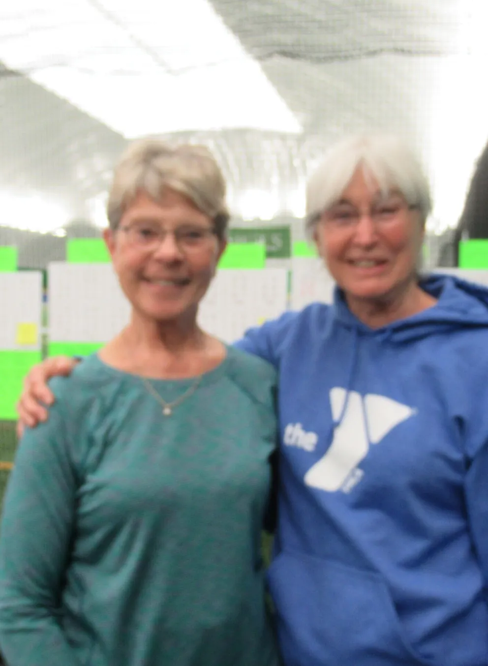 Two women stand together in an indoor sports facility; one wears a teal shirt, the other a blue hoodie.  Green scoreboards are visible in the background.