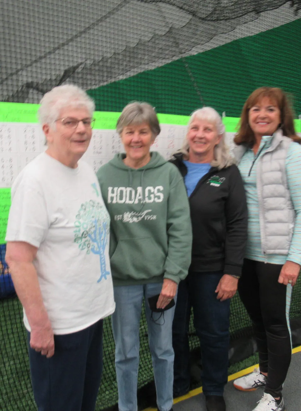 Four women stand smiling near score sheets; one wears a green sweatshirt, another a white shirt, and two others have on darker jackets.  They're in an indoor sports facility.