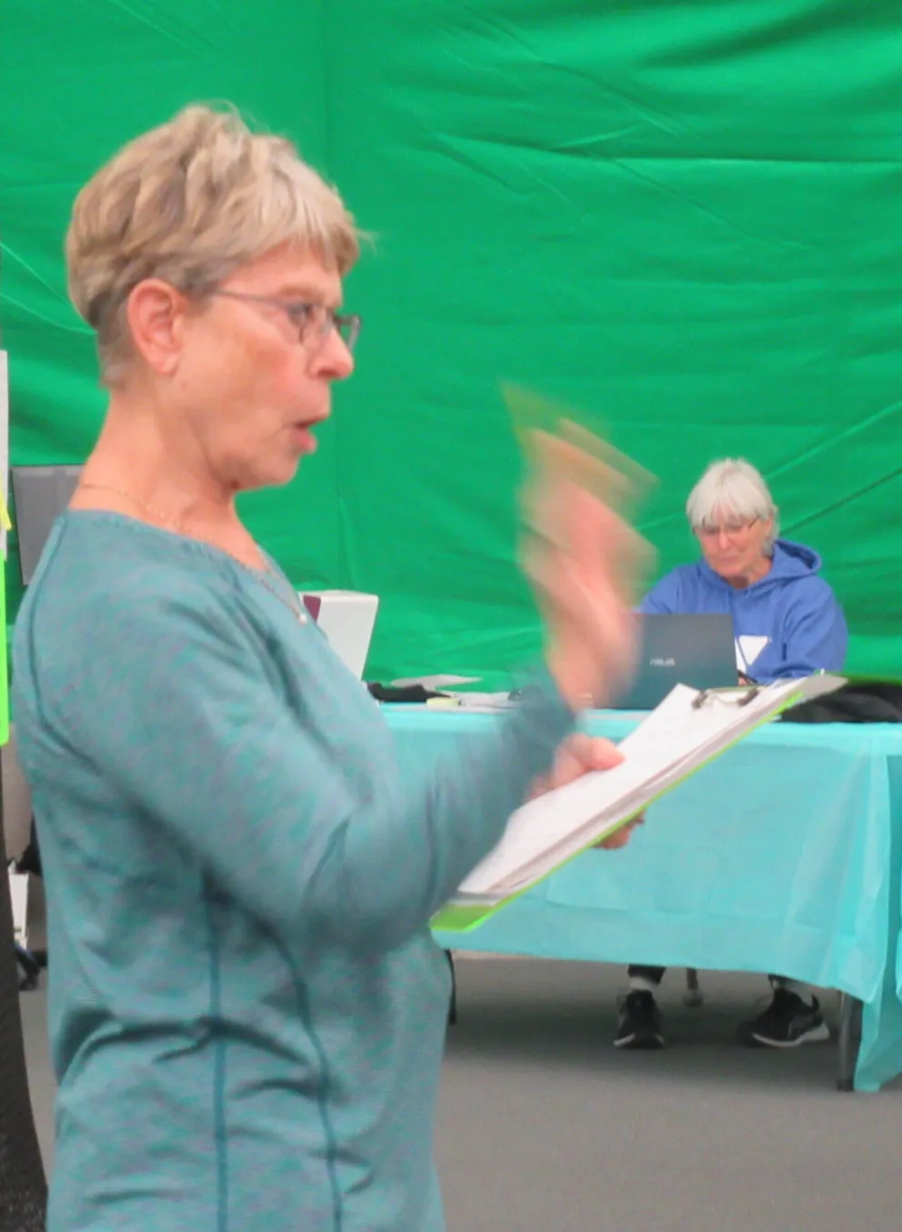 A woman in teal, holding a clipboard, addresses a group.  Bright green backdrop.  Others are seated at a light blue table, some with laptops.  Notes hang on a black mesh screen.