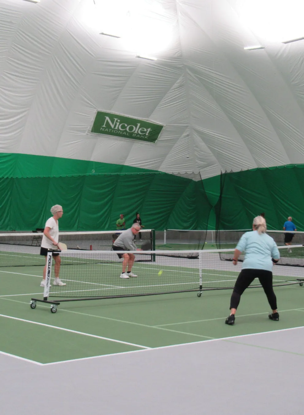 Active adults playing pickleball indoors on several courts.  Nicolet Athletic Center banner visible.