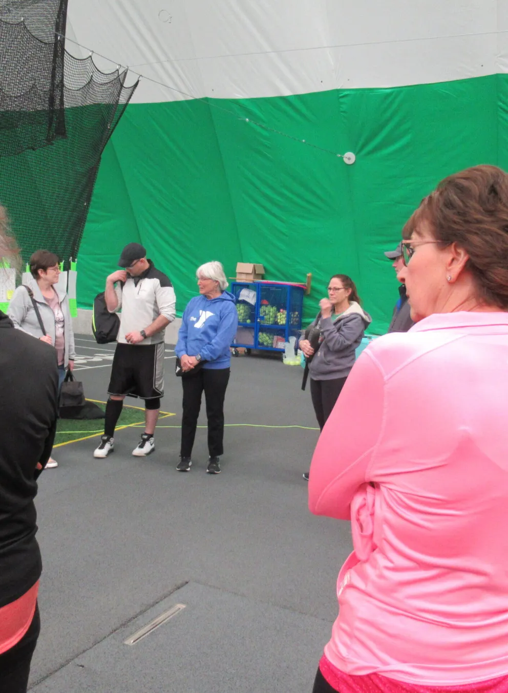 A group of adults stand in an indoor facility, some appearing to listen to instructions.  A green wall is in the background.