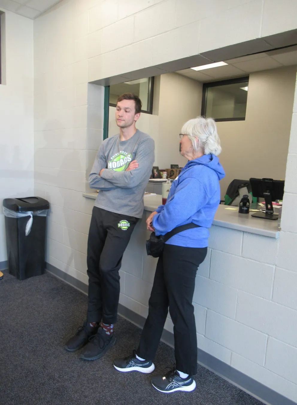 A young man and older woman stand at a counter, talking.  He wears a gray sweatshirt and black sweatpants; she's in a blue jacket and black pants.