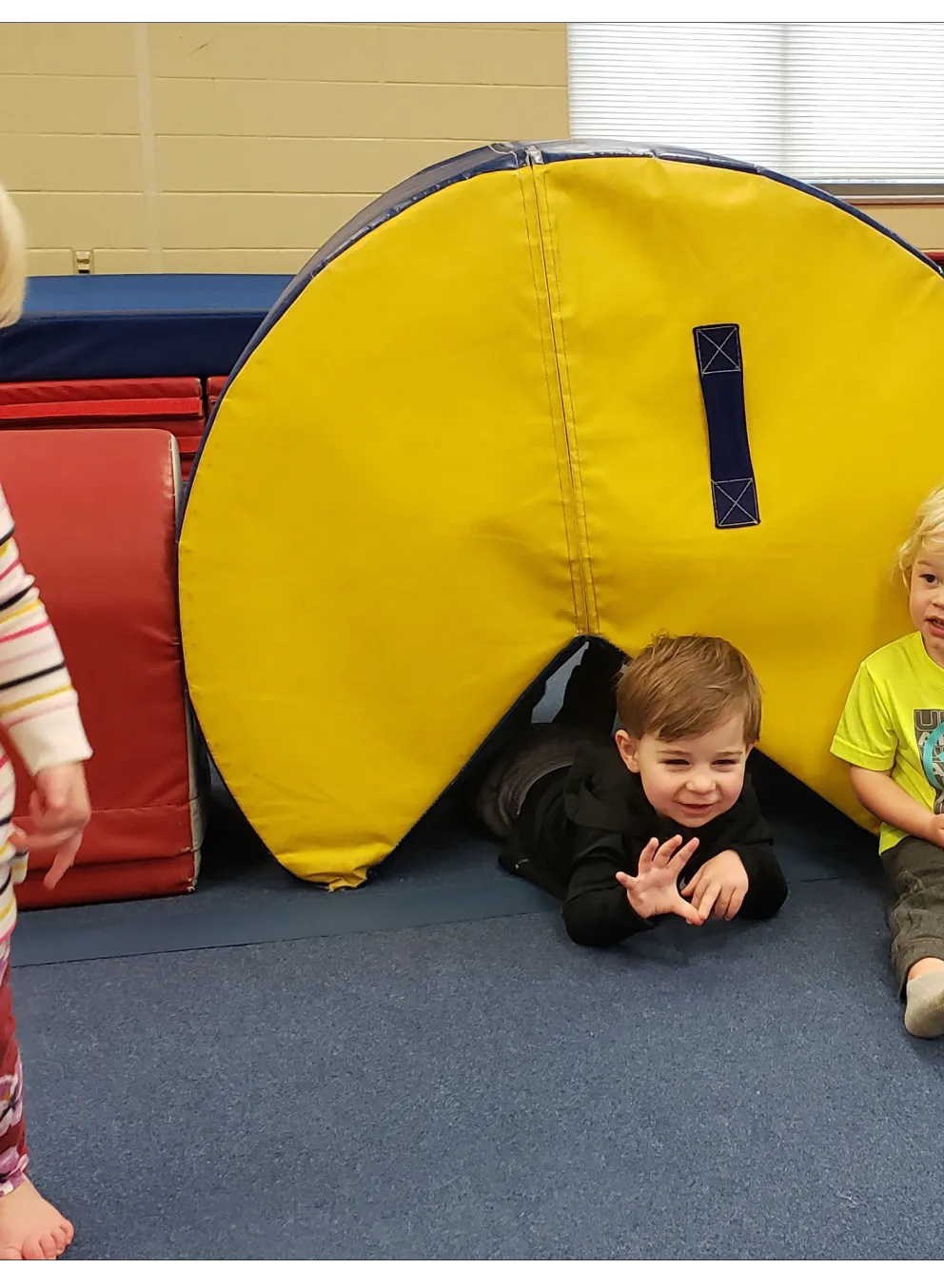 Three toddlers playing in a gym; two boys are near a yellow play tunnel, and a girl stands nearby.