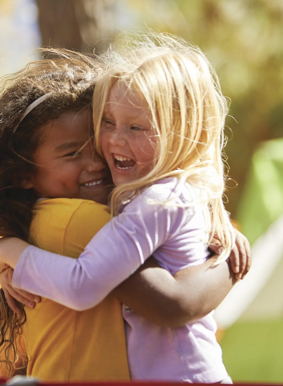 Two girls hug joyfully at a summer camp,  one in a yellow shirt and the other in lavender.