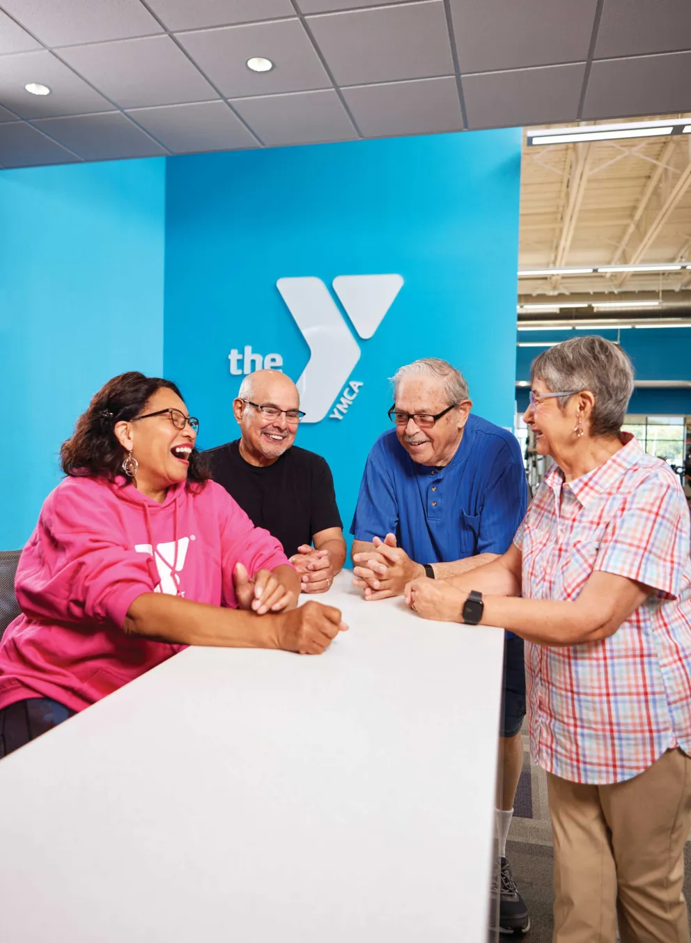Four adults laugh together at a long white table against a bright blue wall with a logo.