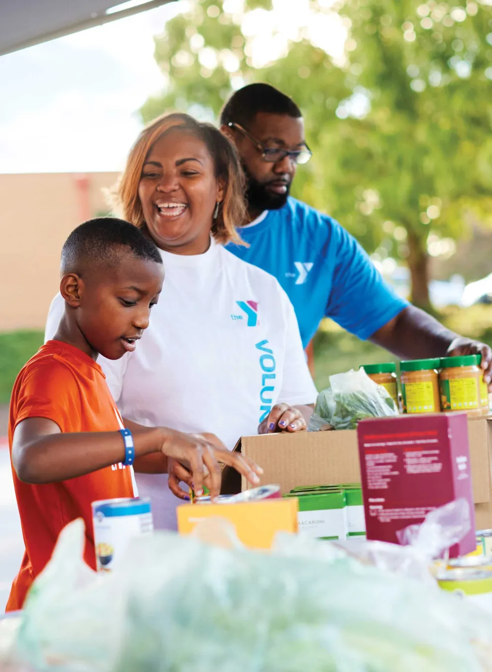 Smiling volunteers, including a boy, sort food donations into boxes.