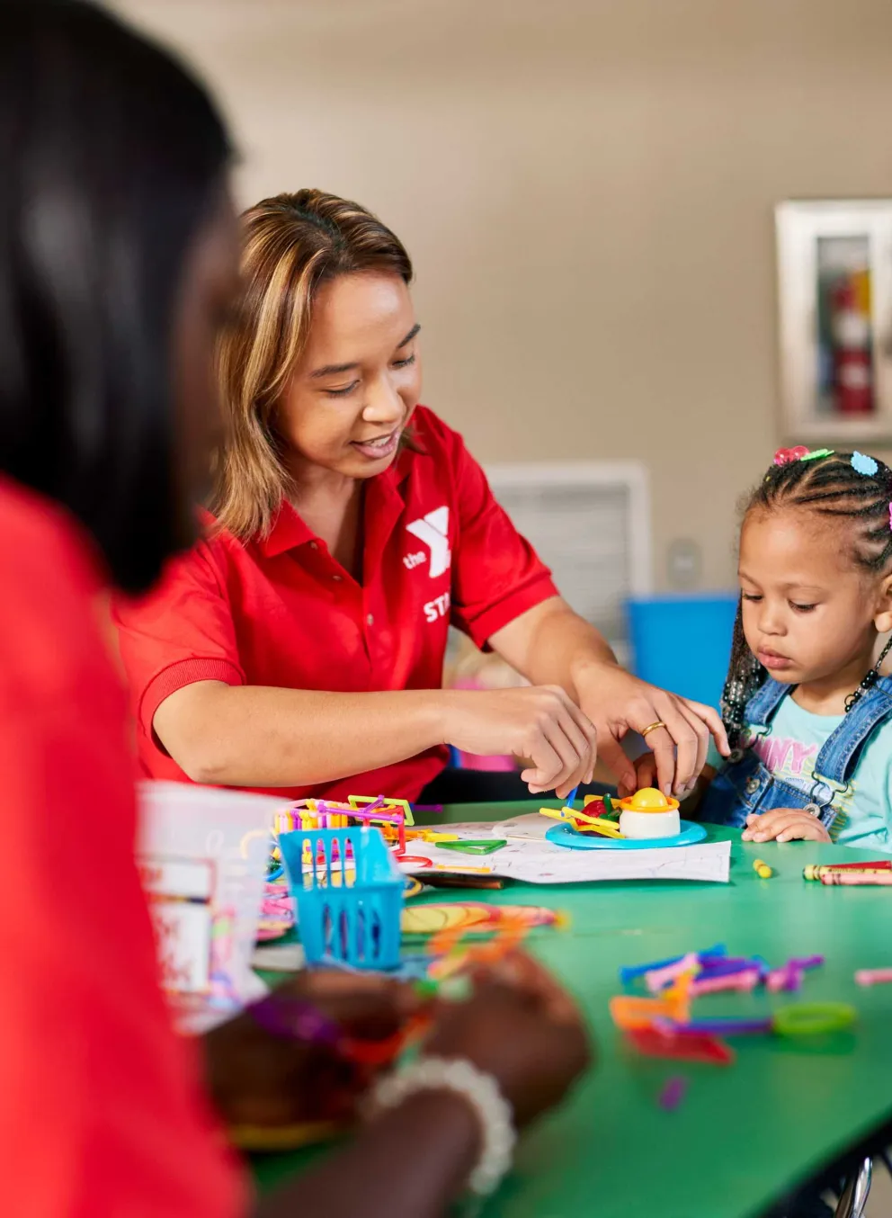 A female counselor assists a young girl with a craft project at a kids' activity table.  Another child sits nearby, and a second adult is visible in the background. Colorful art supplies are scattered on the table.