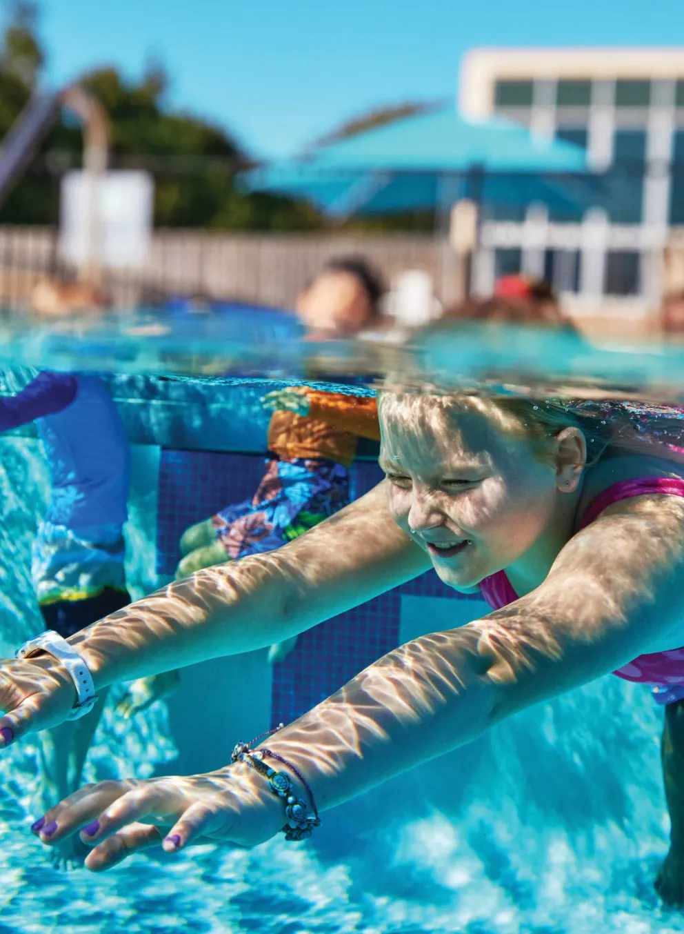 A girl in a pink swimsuit swims underwater in a pool; other children are visible in the background.