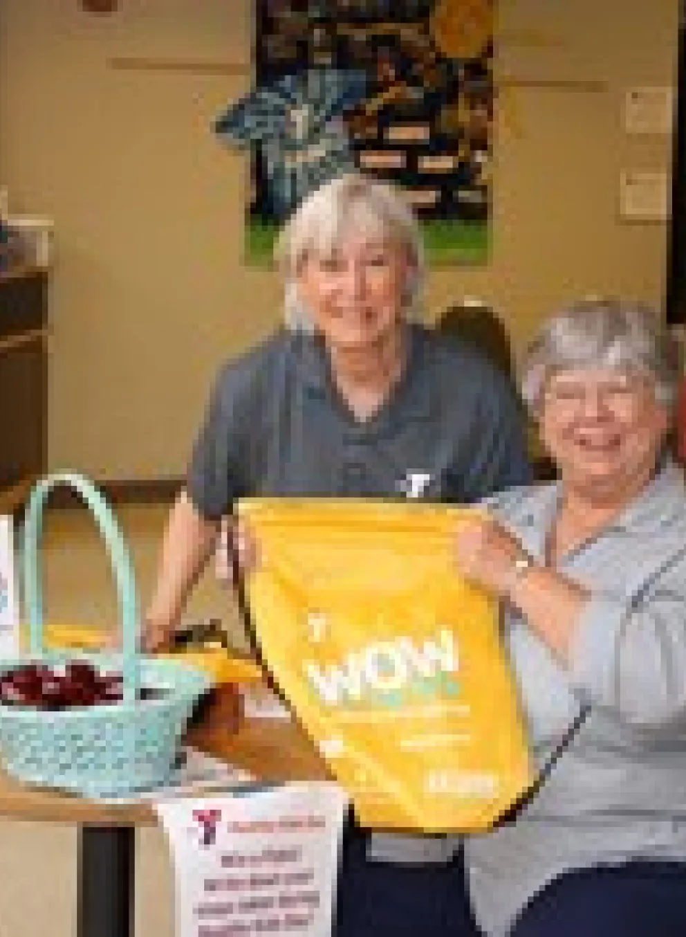 Two smiling women stand behind a table filled with treats and information at a community center event. The lady holds a yellow tote bag labeled "WOW".