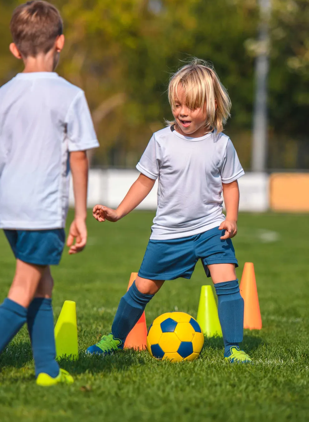 Kids in white soccer jerseys and navy shorts practice dribbling a soccer ball around cones on a grassy field.