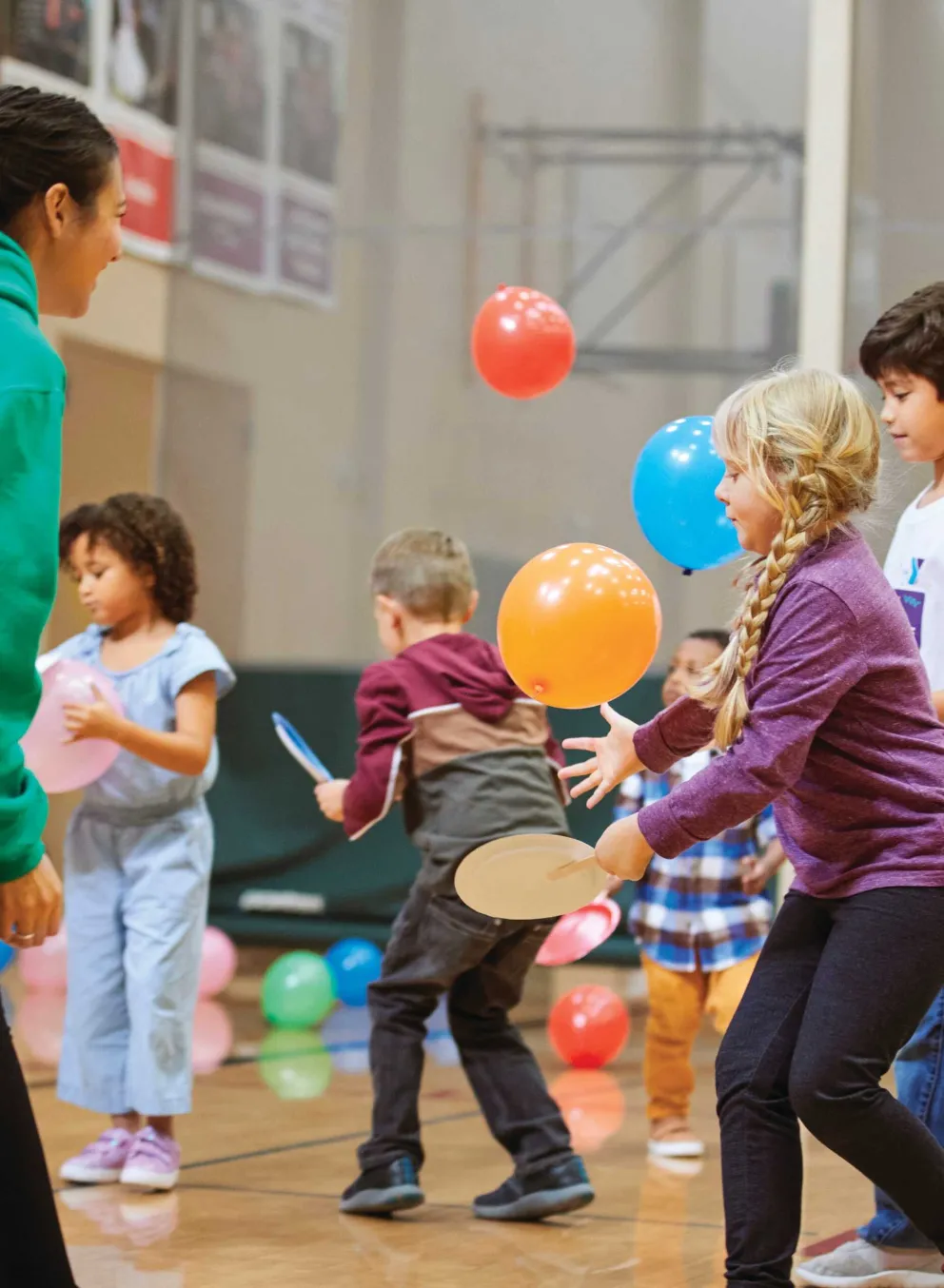 Kids play balloon games in a gym with a counselor.  Colorful balloons float, children use paddles, and smiles abound.