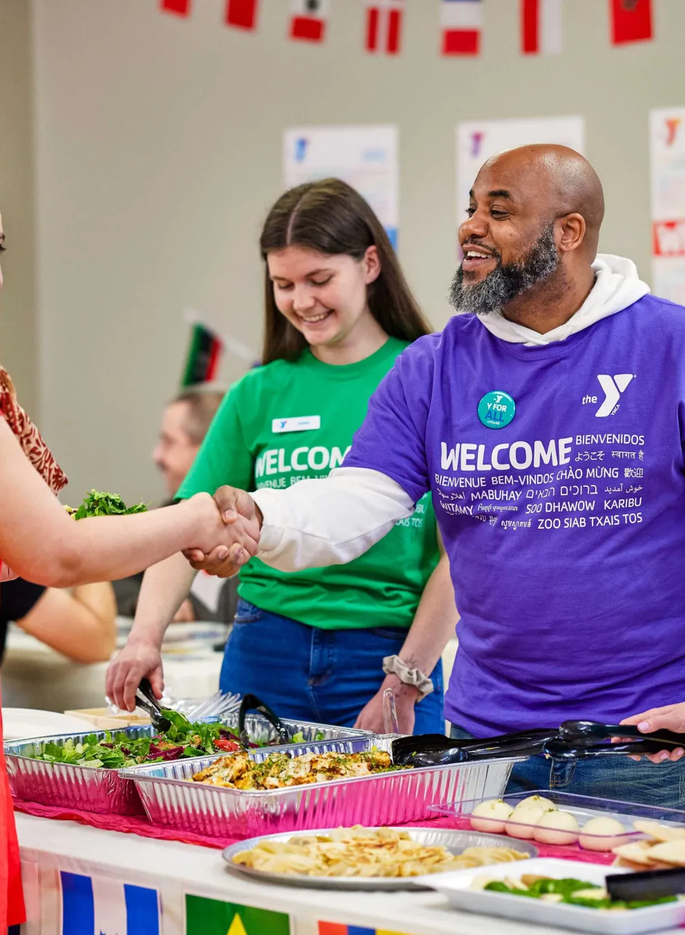 Smiling people shake hands at a community event; food is on tables decorated with international flags.