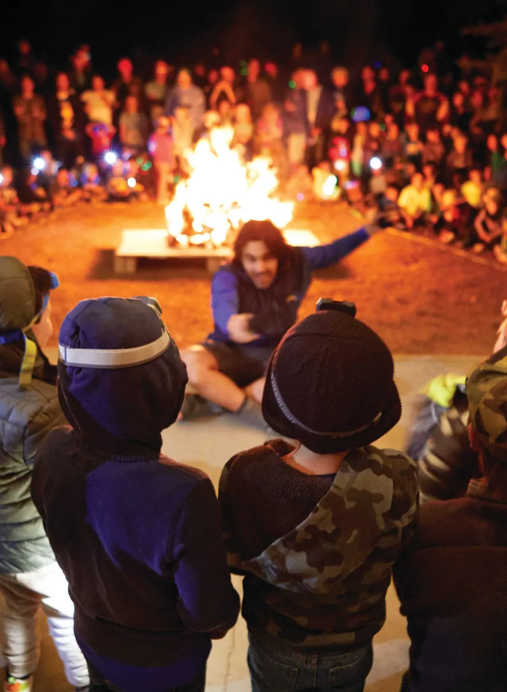 Children gather around a bonfire, captivated by a storyteller.  A large crowd watches from behind the kids. Warm light illuminates the scene.