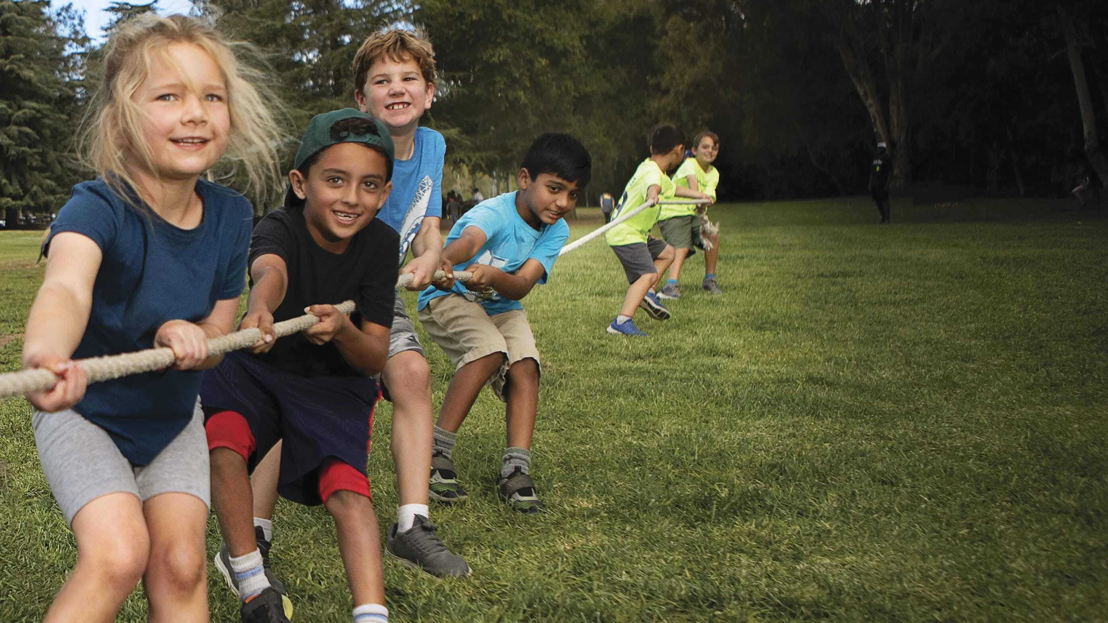 Kids joyfully play tug-of-war on a grassy field, under a bright blue sky.  Smiling children pull a thick rope, their faces full of energy and excitement.