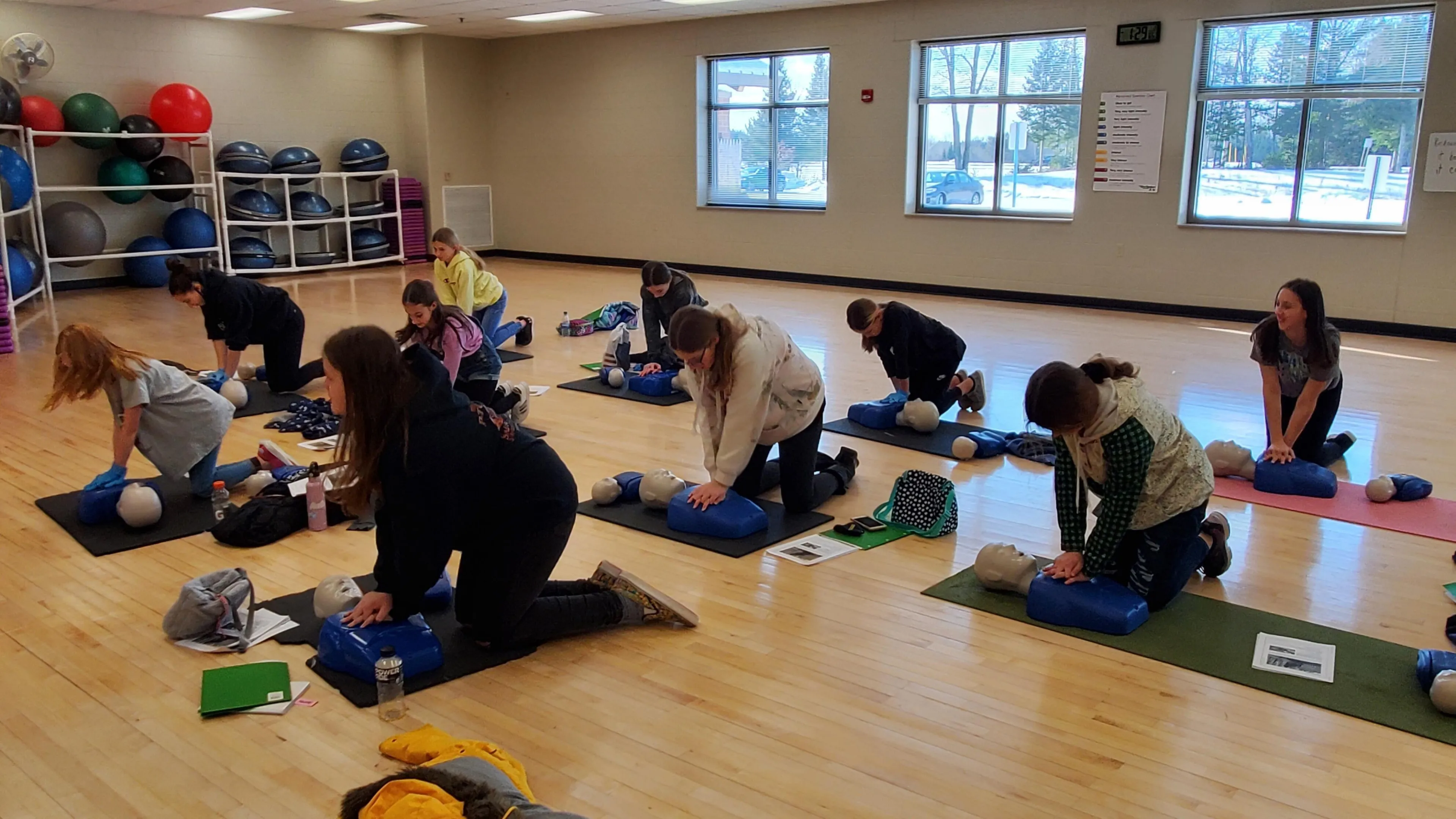 Several young adults kneel on mats, practicing CPR on training dummies in a gym.  Exercise balls are visible in the background.