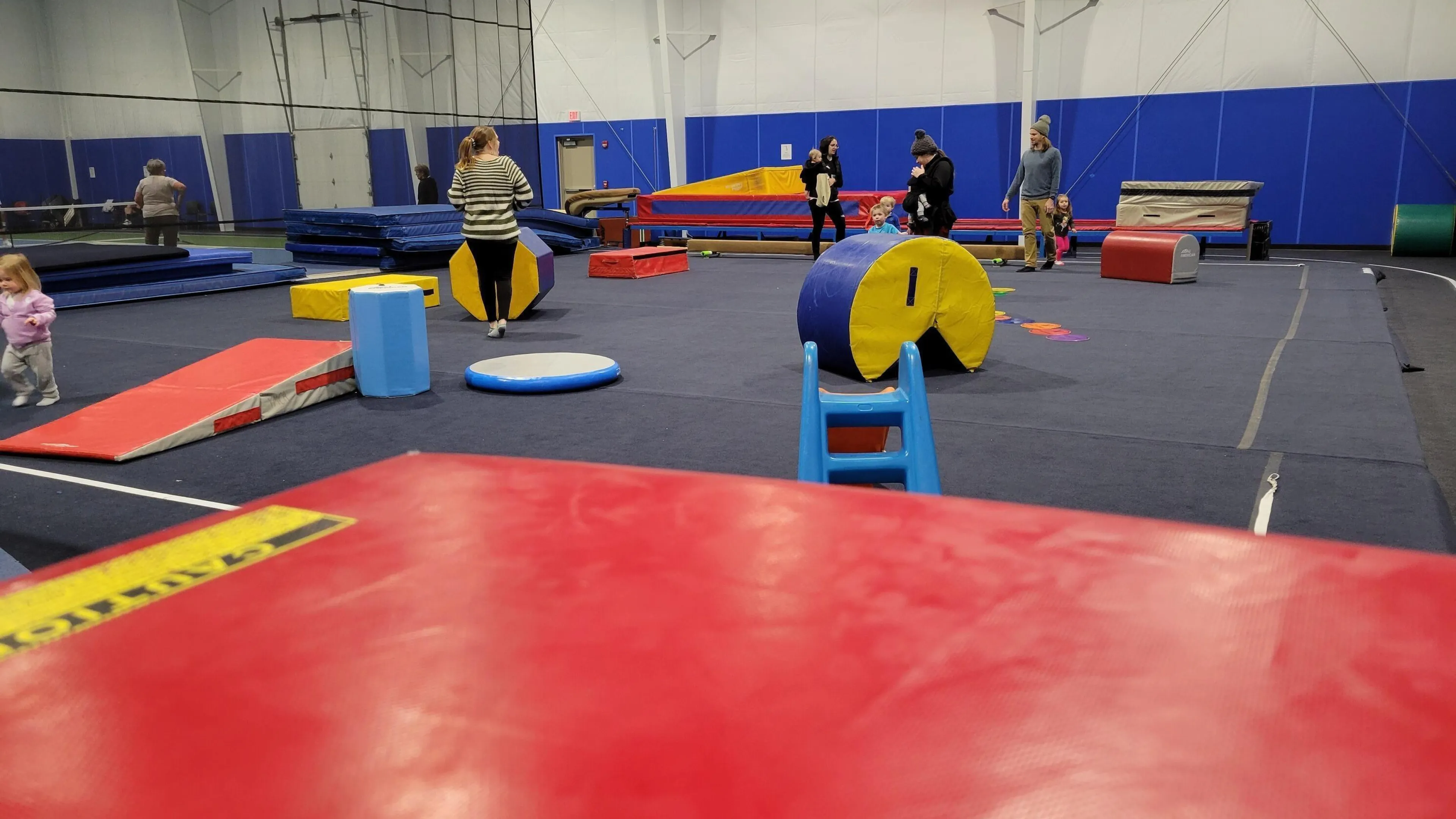 Kids' gymnastics class in session.  Toddlers and adults are present. Colorful mats, blocks, and other equipment are scattered around a large gym floor. A little girl is playing near some mats.