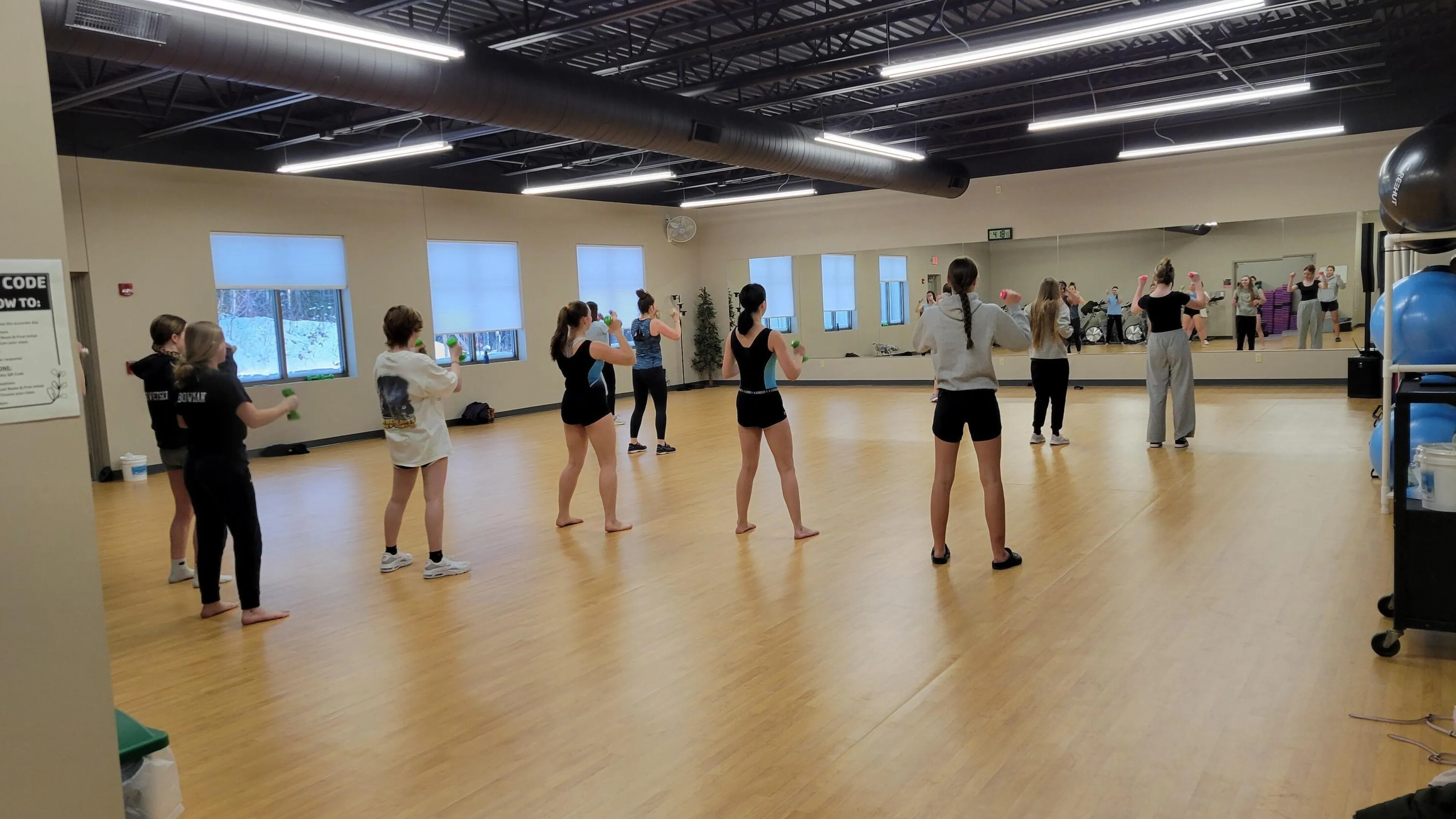 Young women in athletic wear participate in a fitness class, some holding light weights, in a bright, spacious room with wood floors and large windows.