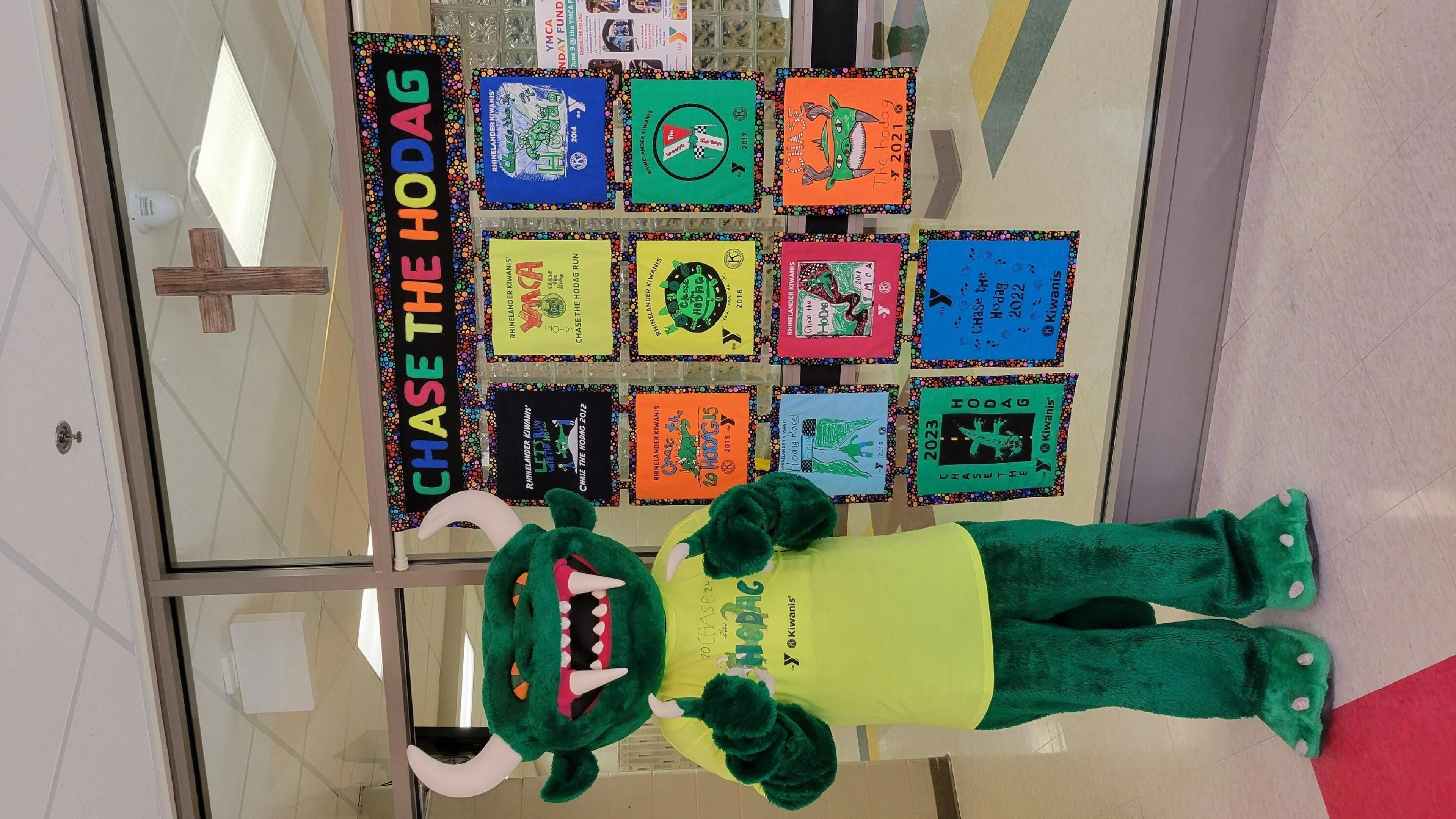 Green Hodag mascot in lime green shirt stands by colorful event posters;  "Chase the Hodag" banner above.