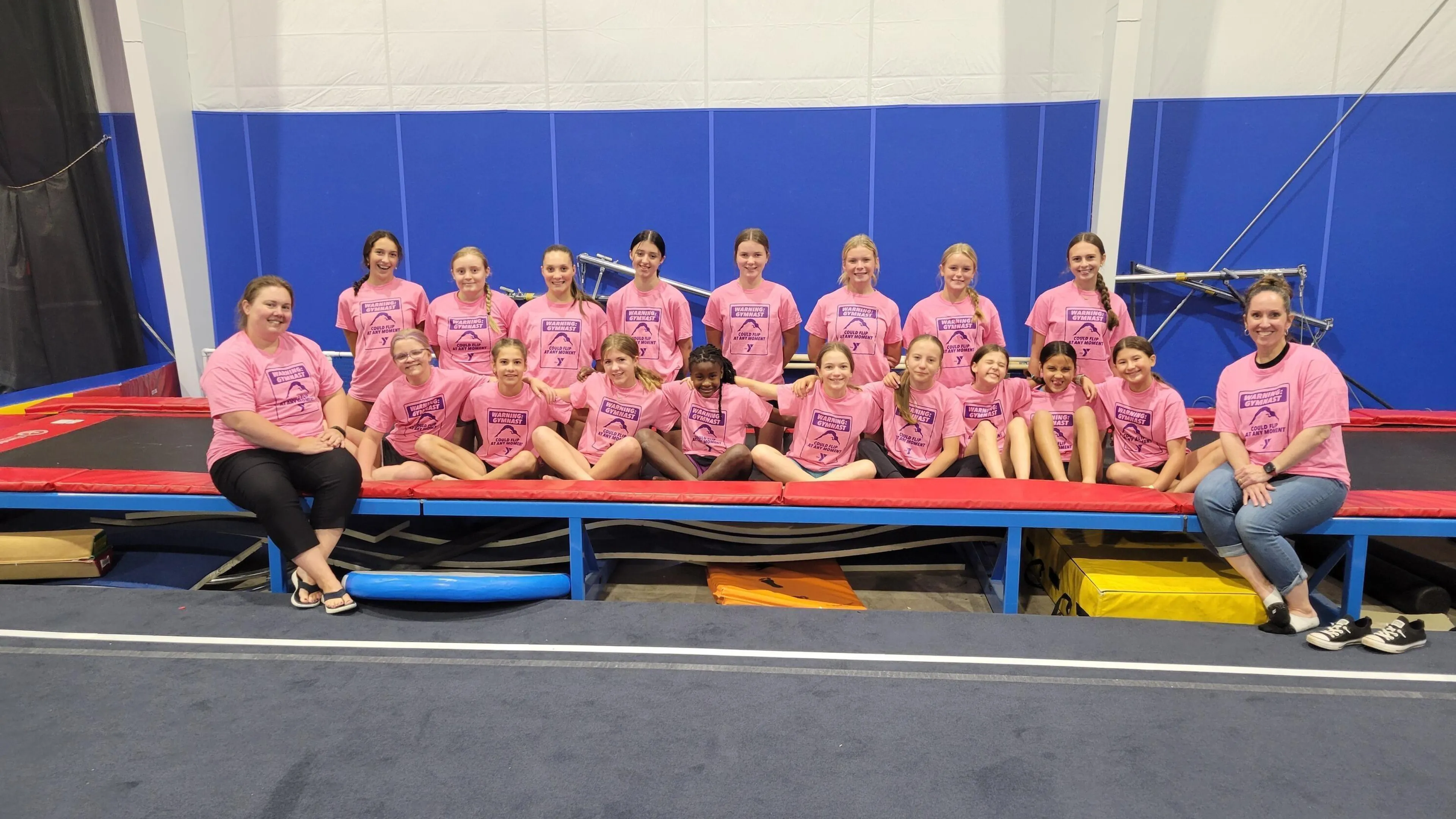 A group of girls in pink t-shirts sits on a padded gymnastics platform, two adult women flank them.  A trampoline is behind them.