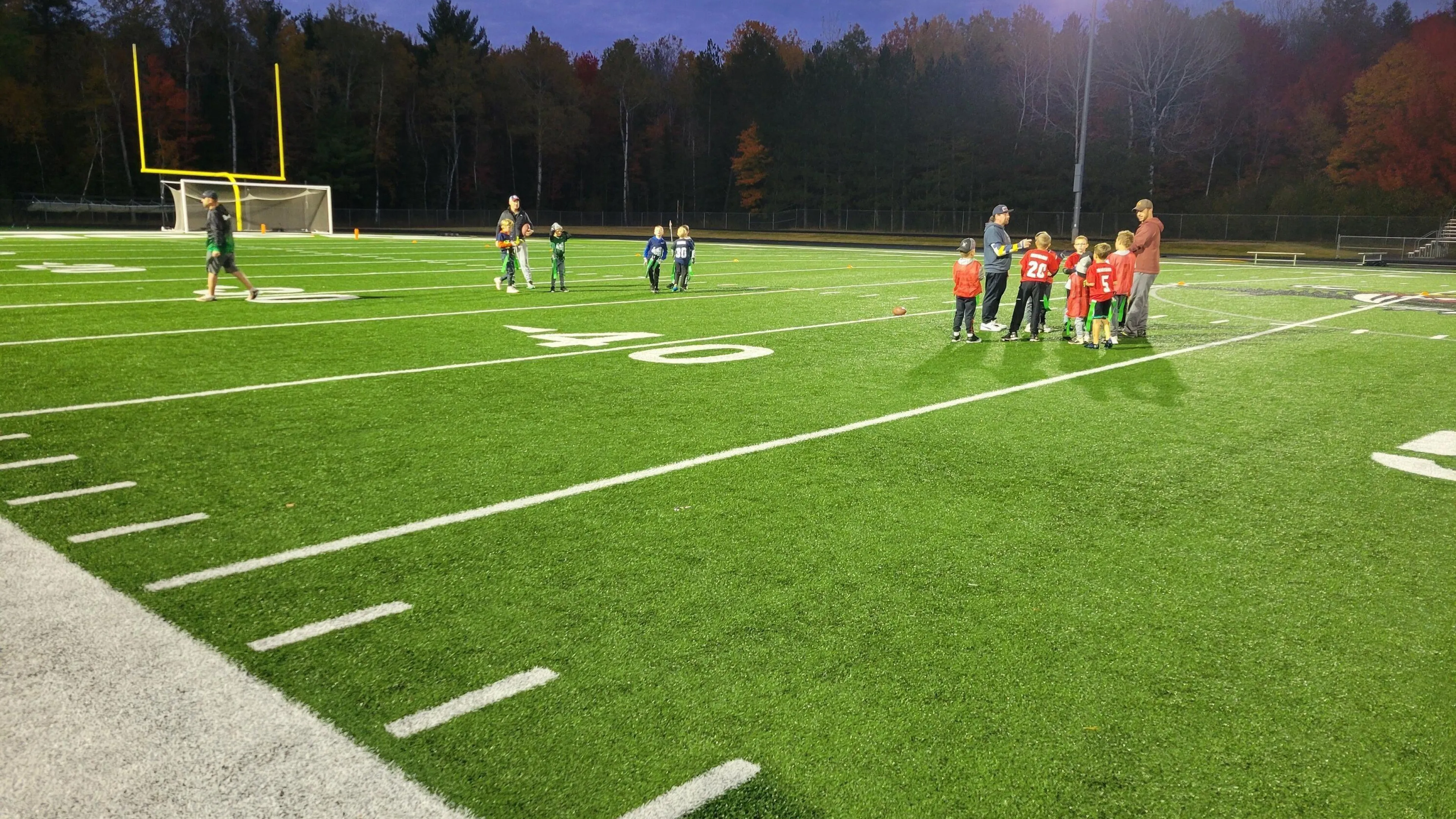 Kids in red jerseys participate in a football practice on a well-lit field, with coaches providing instruction under the evening sky.  A few other kids and adults are nearby.
