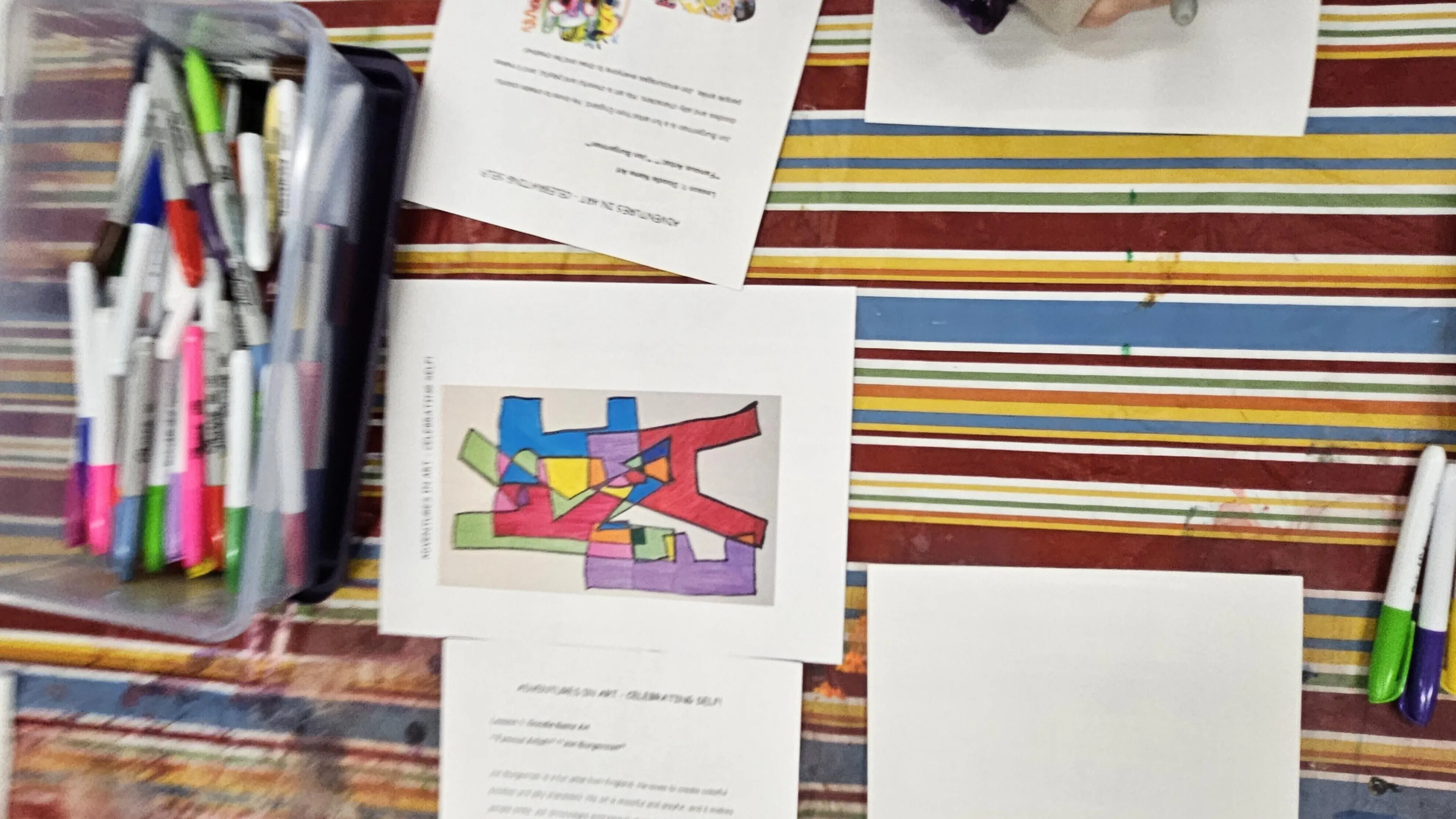 Colorful markers in a container, papers with instructions and a child's abstract art piece on a striped table.