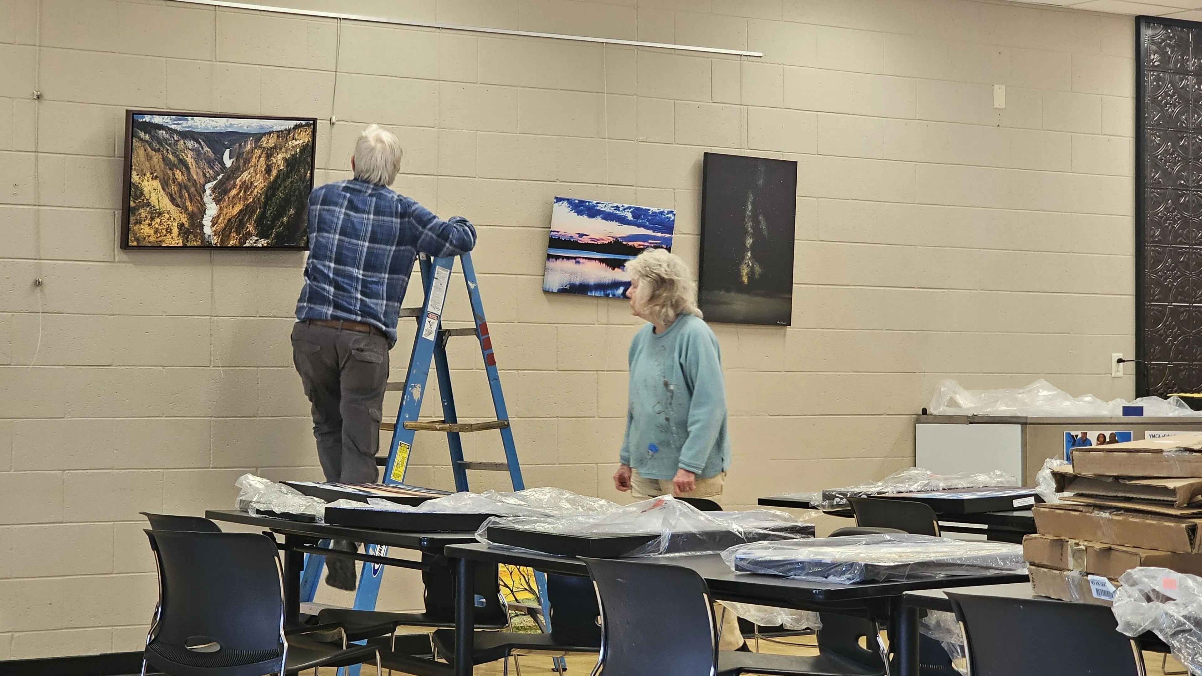 Two adults hang artwork on a wall; one uses a stepladder.  Several framed landscape photos are visible, along with tables covered in plastic-wrapped items.