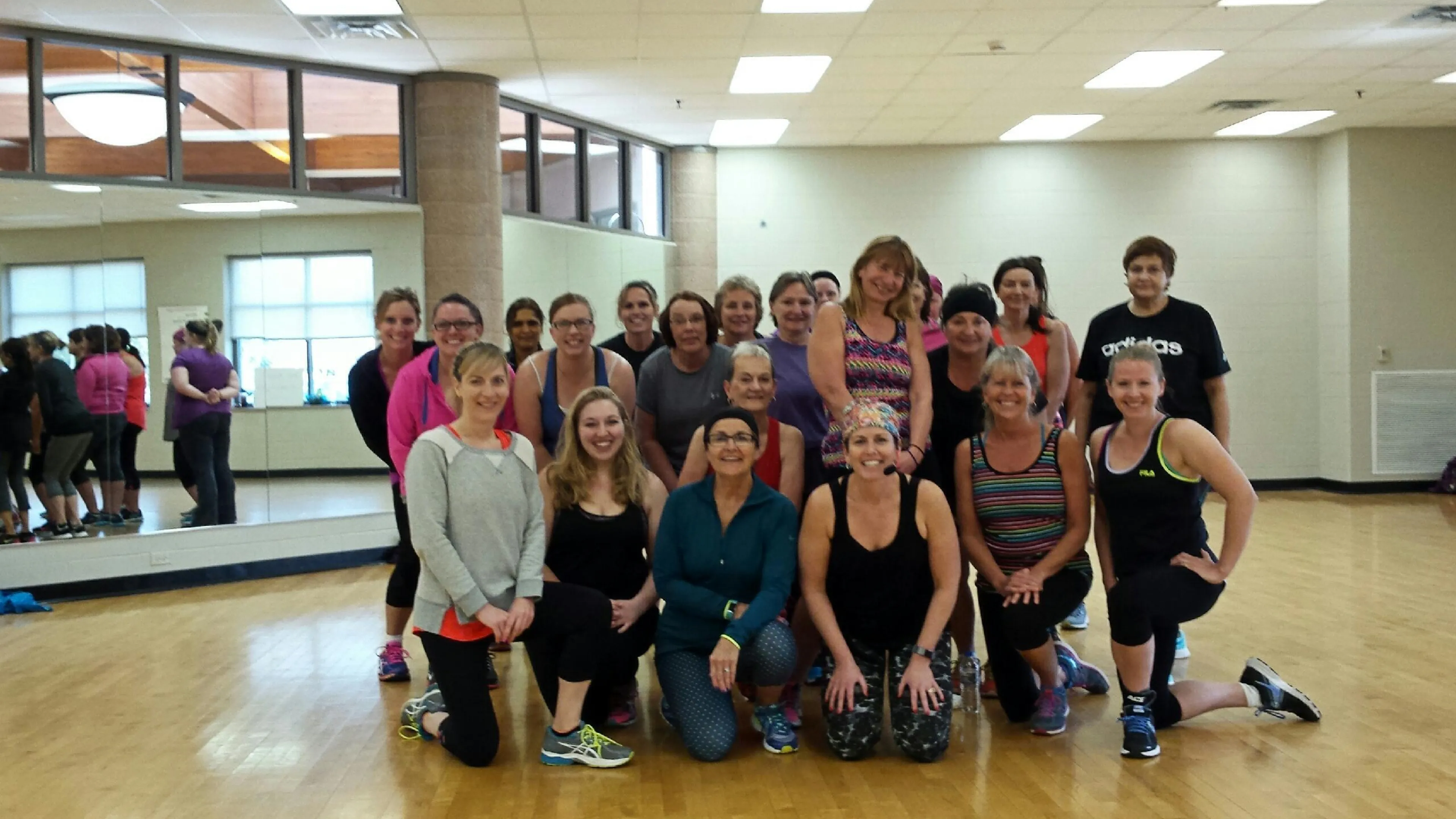 A large group of women and one man pose in a gym; many are kneeling.  They appear to be participating in a fitness class.